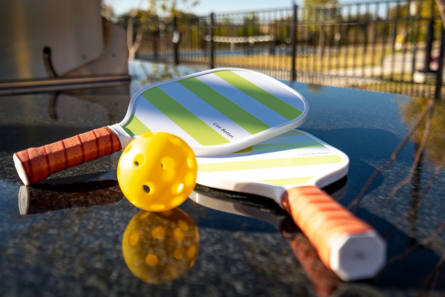Two pickleball paddles and a yellow ball on a black surface outdoors, with a blurred background.