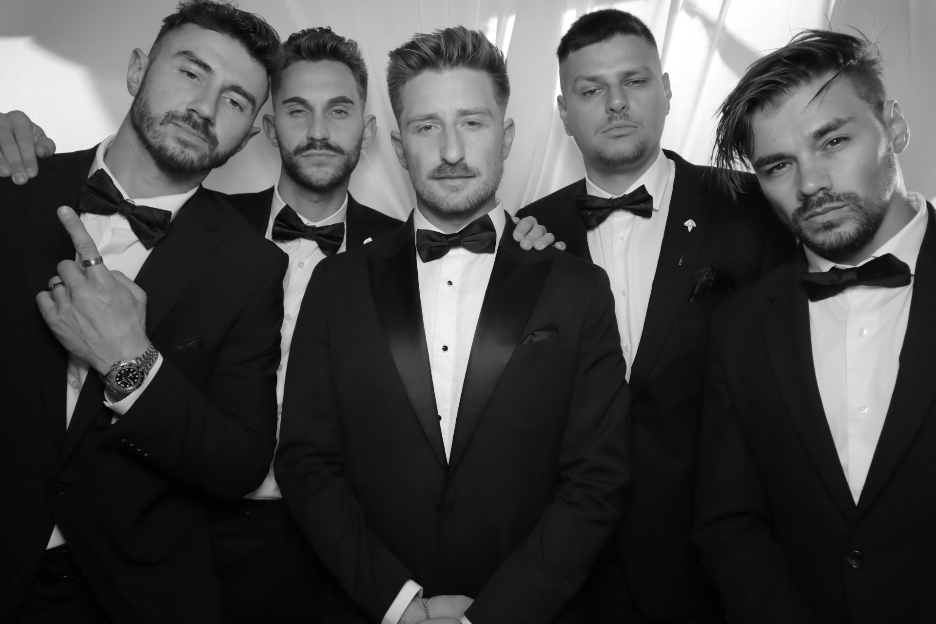 Five men in tuxedos posing together against a dark background