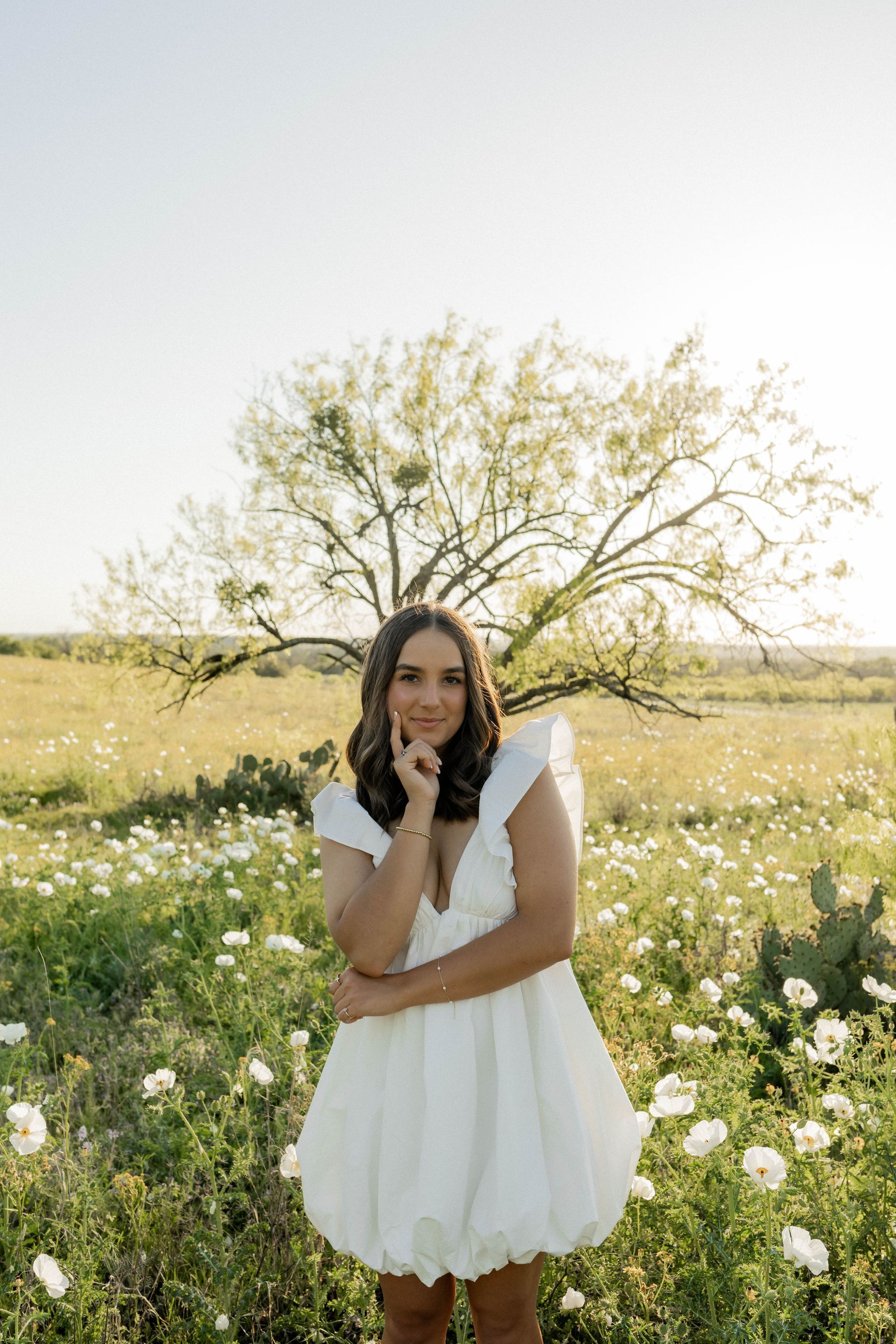 Woman in a white dress standing in a sunlit wildflower field by a tree