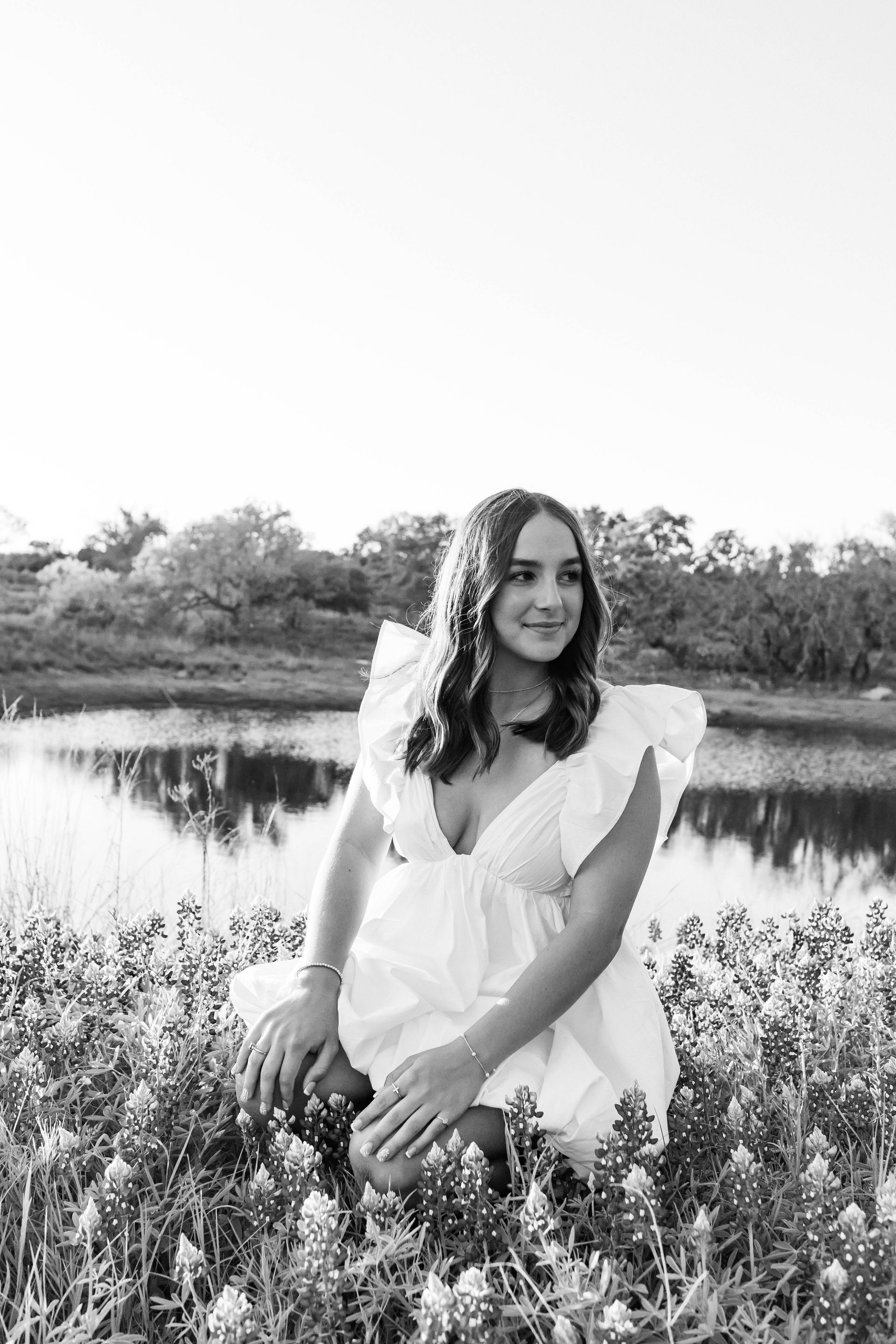 Woman in a white dress standing by a calm lake with trees in the background.
