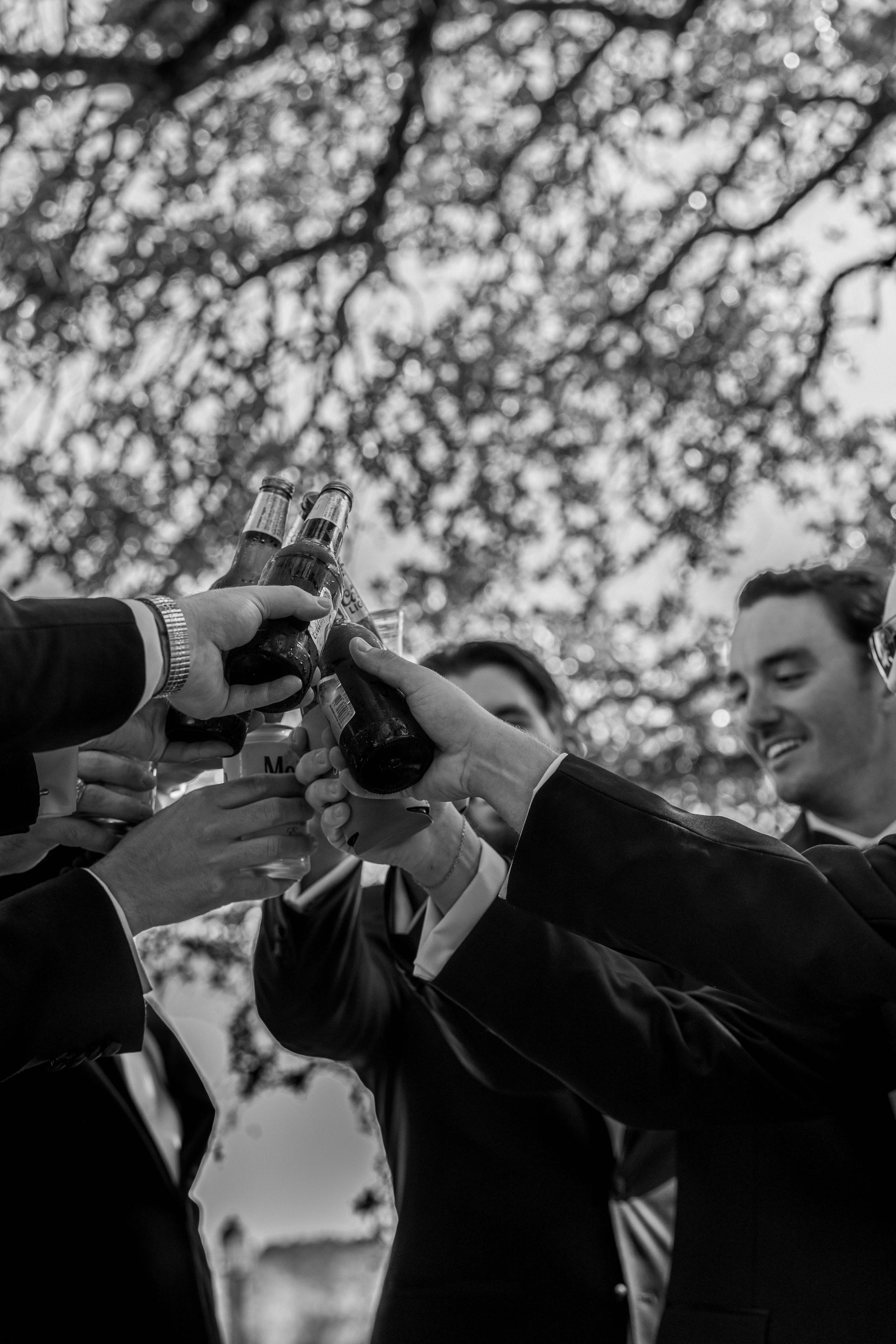 People clinking bottles in a celebratory toast outdoors under tree branches