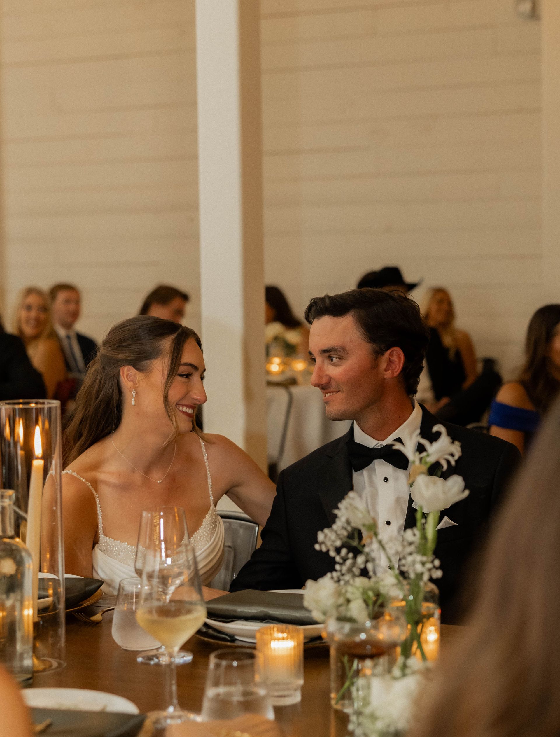 Smiling couple seated at a candlelit dinner reception, looking at each other across the table.