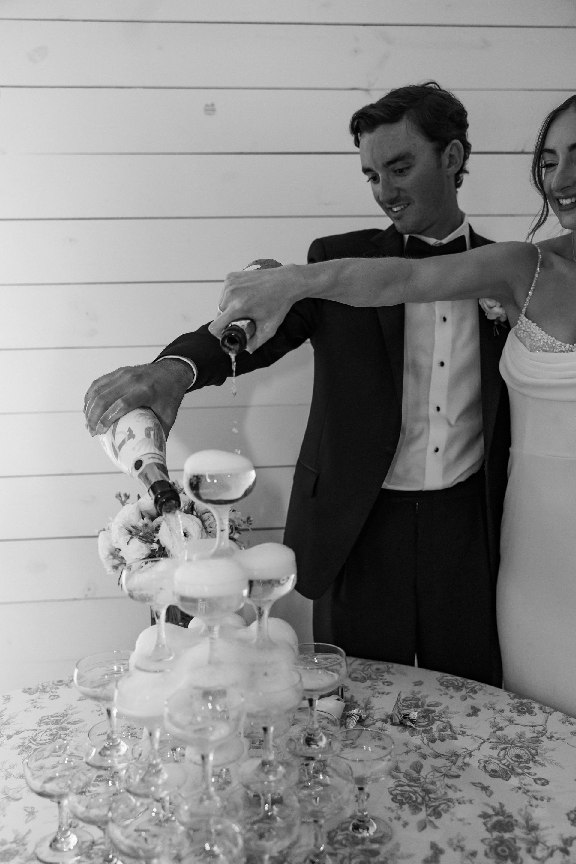 Man and woman in formalwear pouring champagne over a tower of glasses at a decorated table