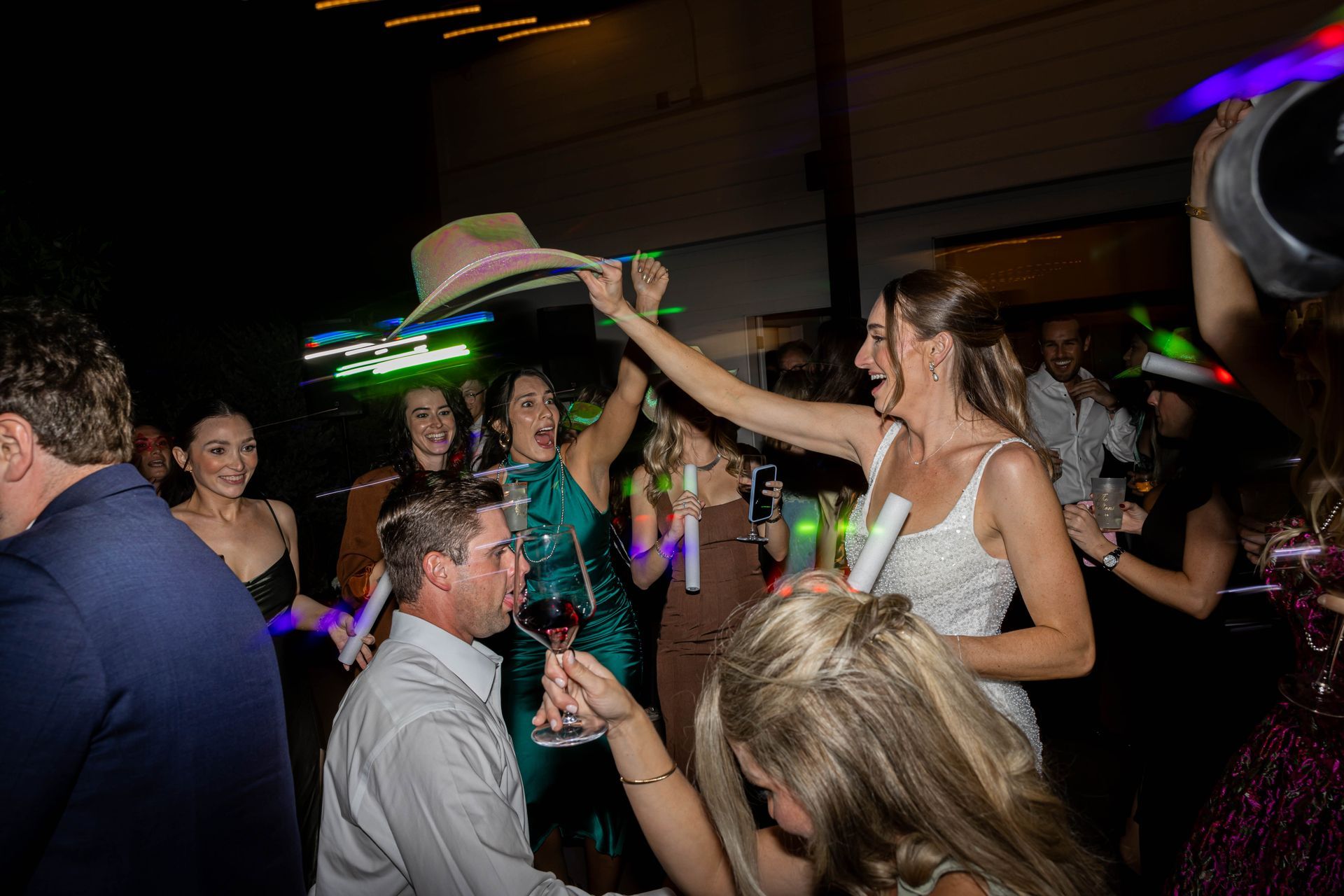 Crowd dancing at a party; woman in white top raises a hat amid colorful lights.