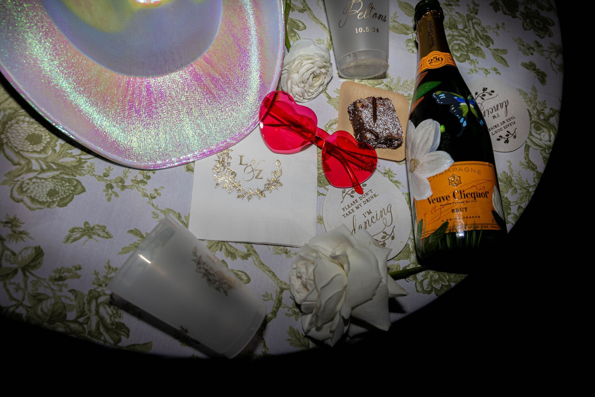 Champagne bottle, red bow, and gift bag on a floral tablecloth under flash lighting