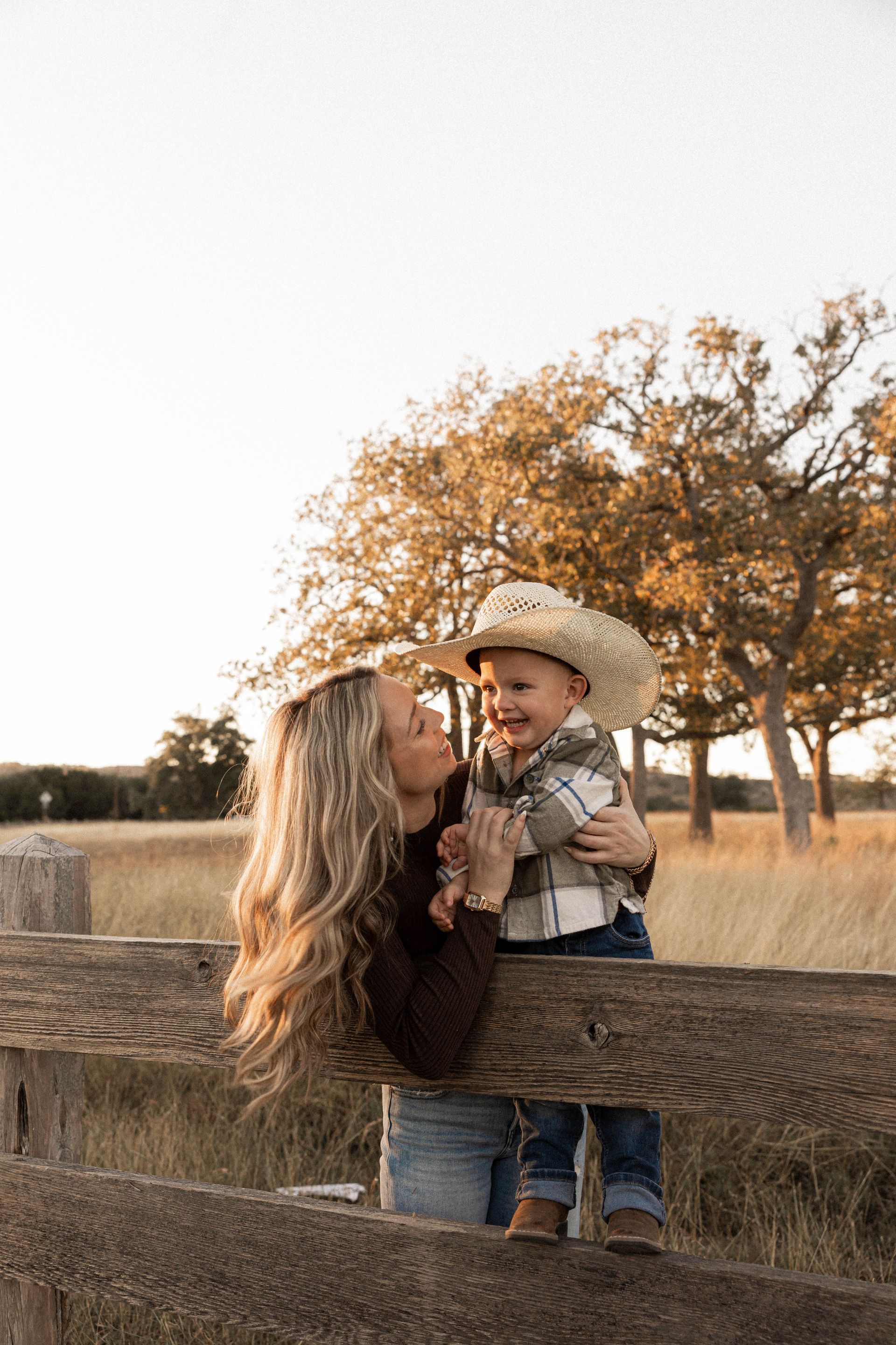 Woman and child by a wooden fence in a field, smiling under autumn trees.