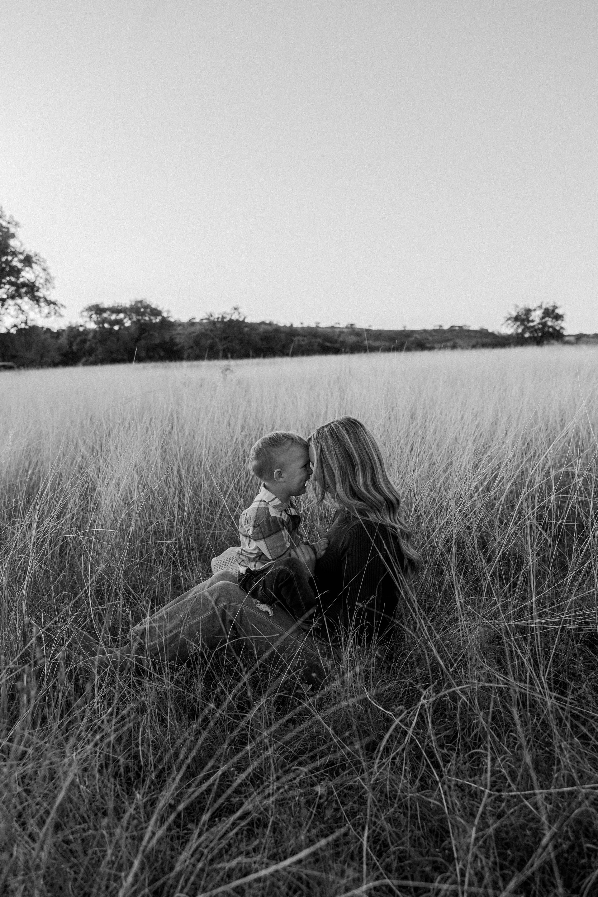 Two people sitting in tall grass in a field, facing away toward a distant tree line.