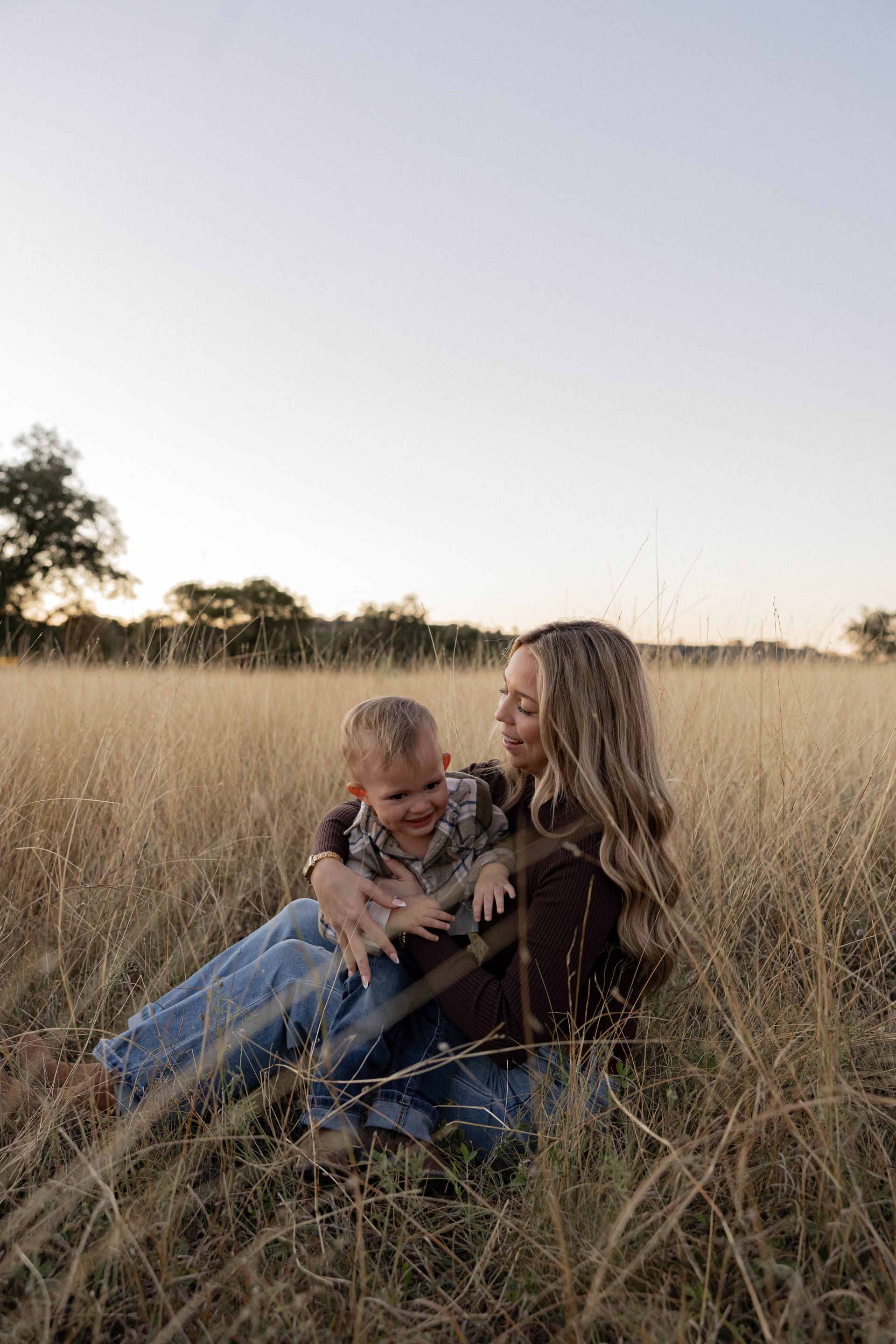 Two people sitting together in tall grass at sunset, smiling and embracing.