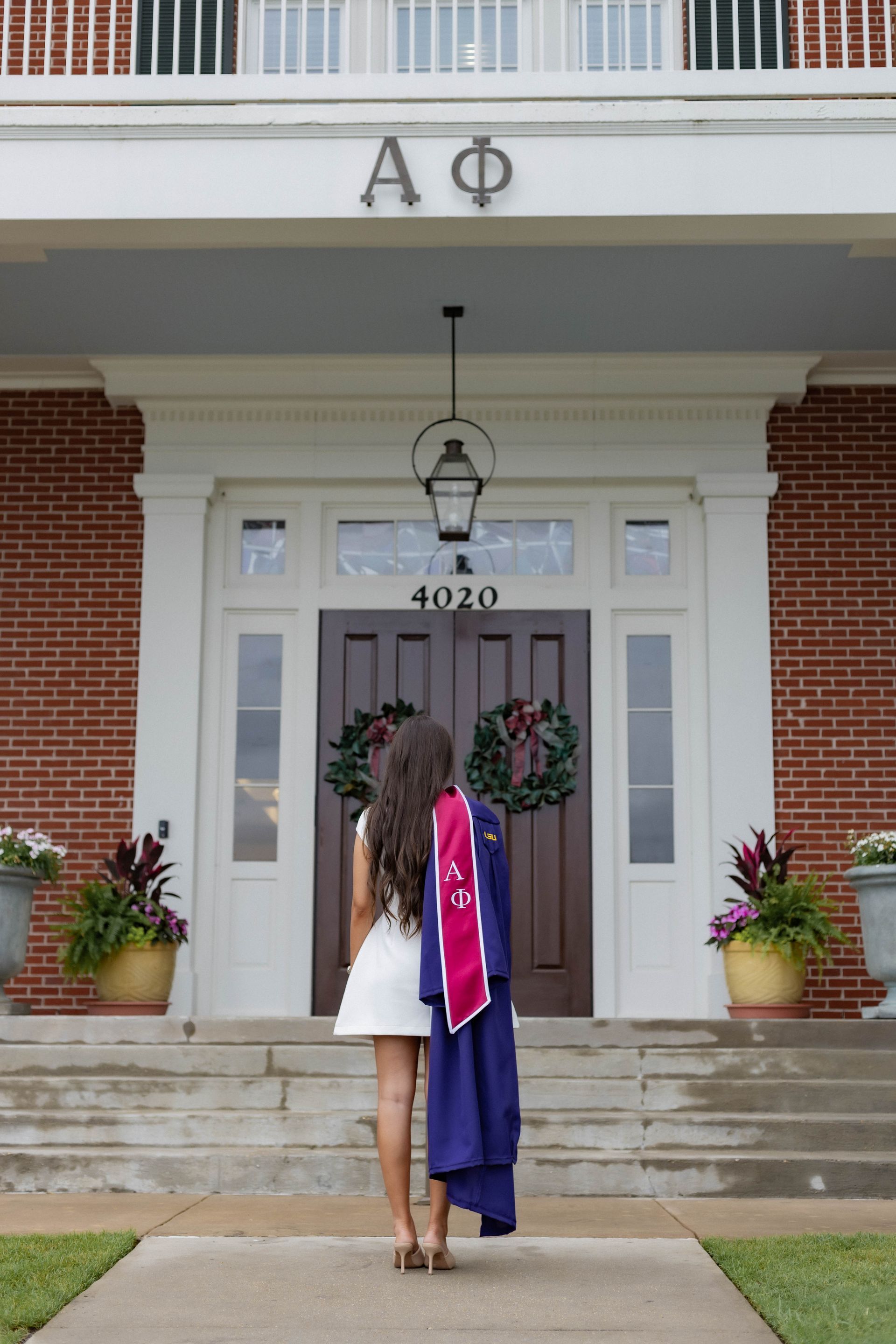 Graduate in purple cap and gown stands with another person at a decorated doorway of a brick building.