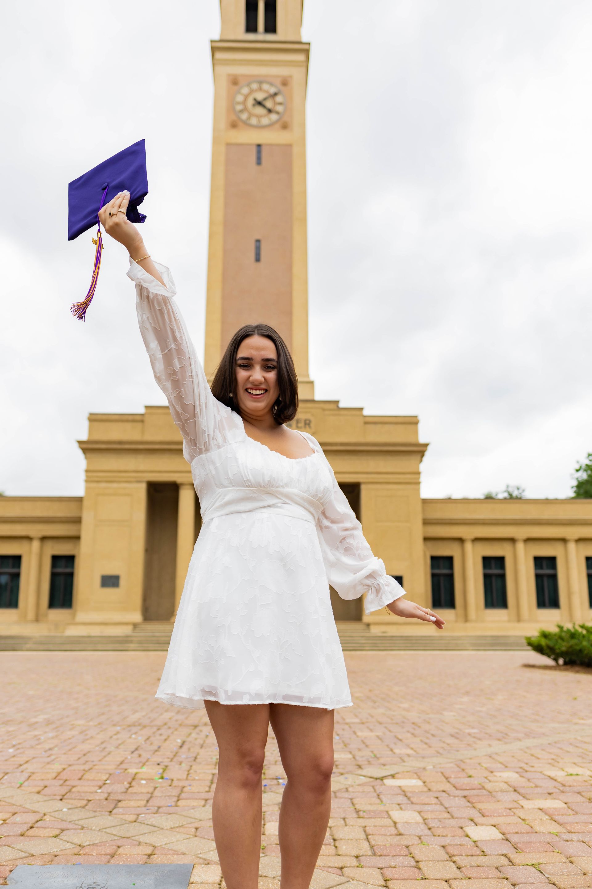 Graduate in white dress holds up a purple cap in front of a clock tower.