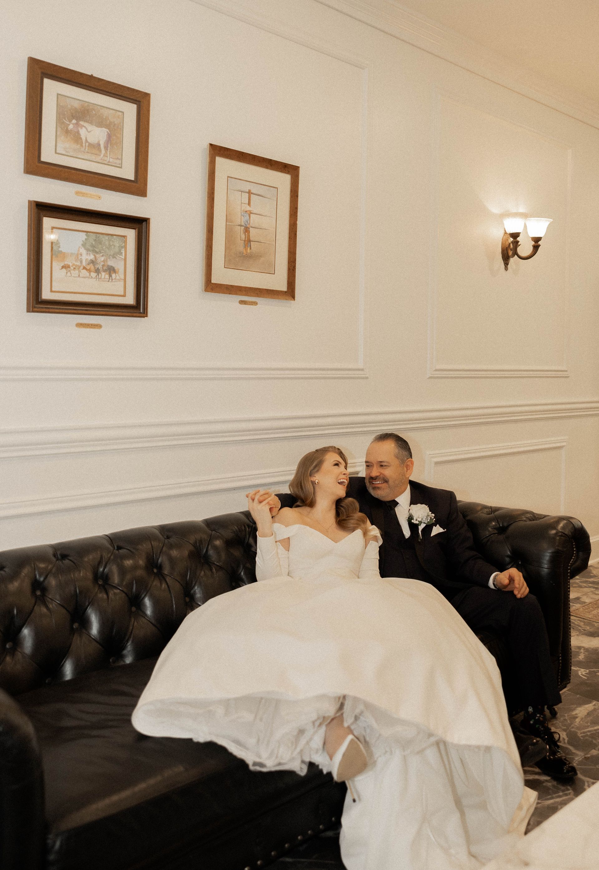 Couple in wedding attire sitting on a black tufted sofa in a softly lit room