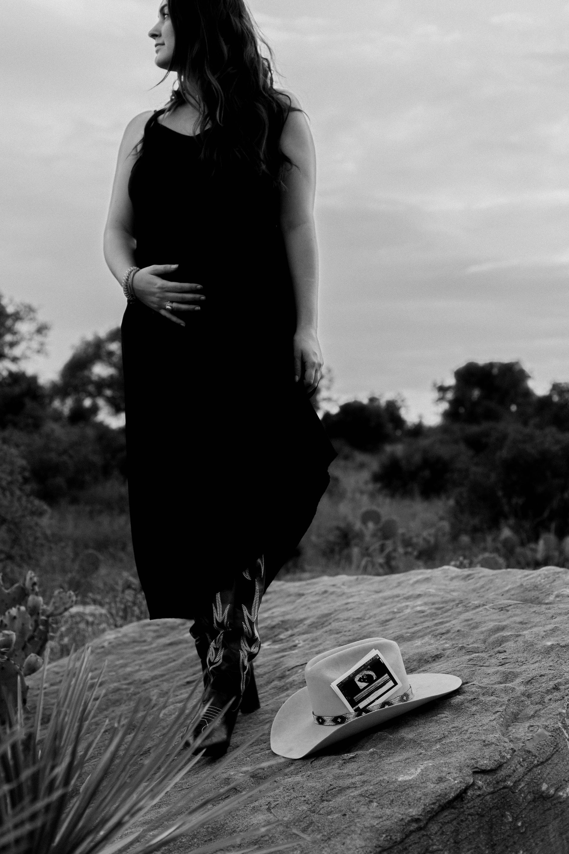 Person in a black dress stands on a rock outdoors beside a hat in a field.