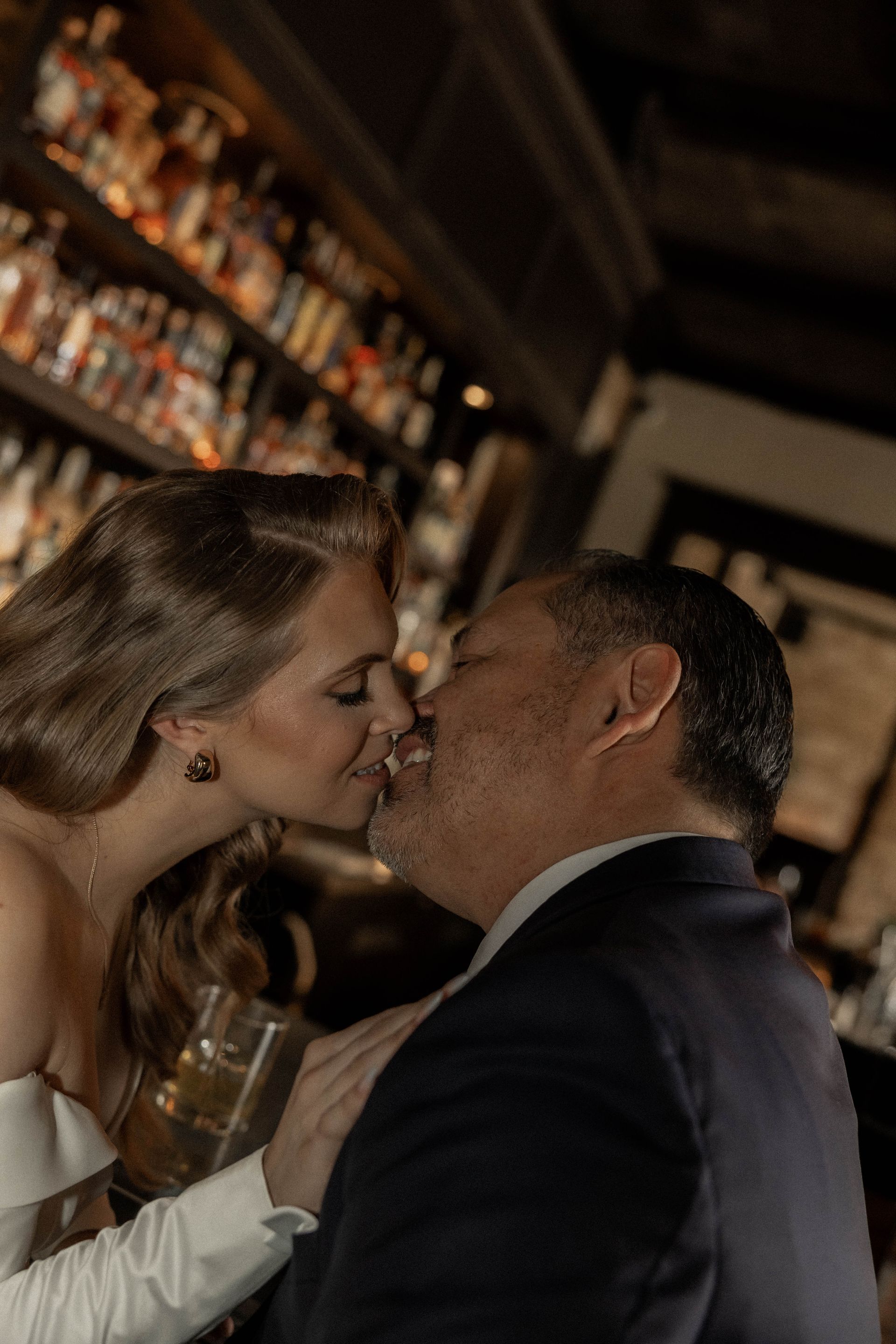 Couple kissing in a dimly lit bar, with shelves of bottles in the background.