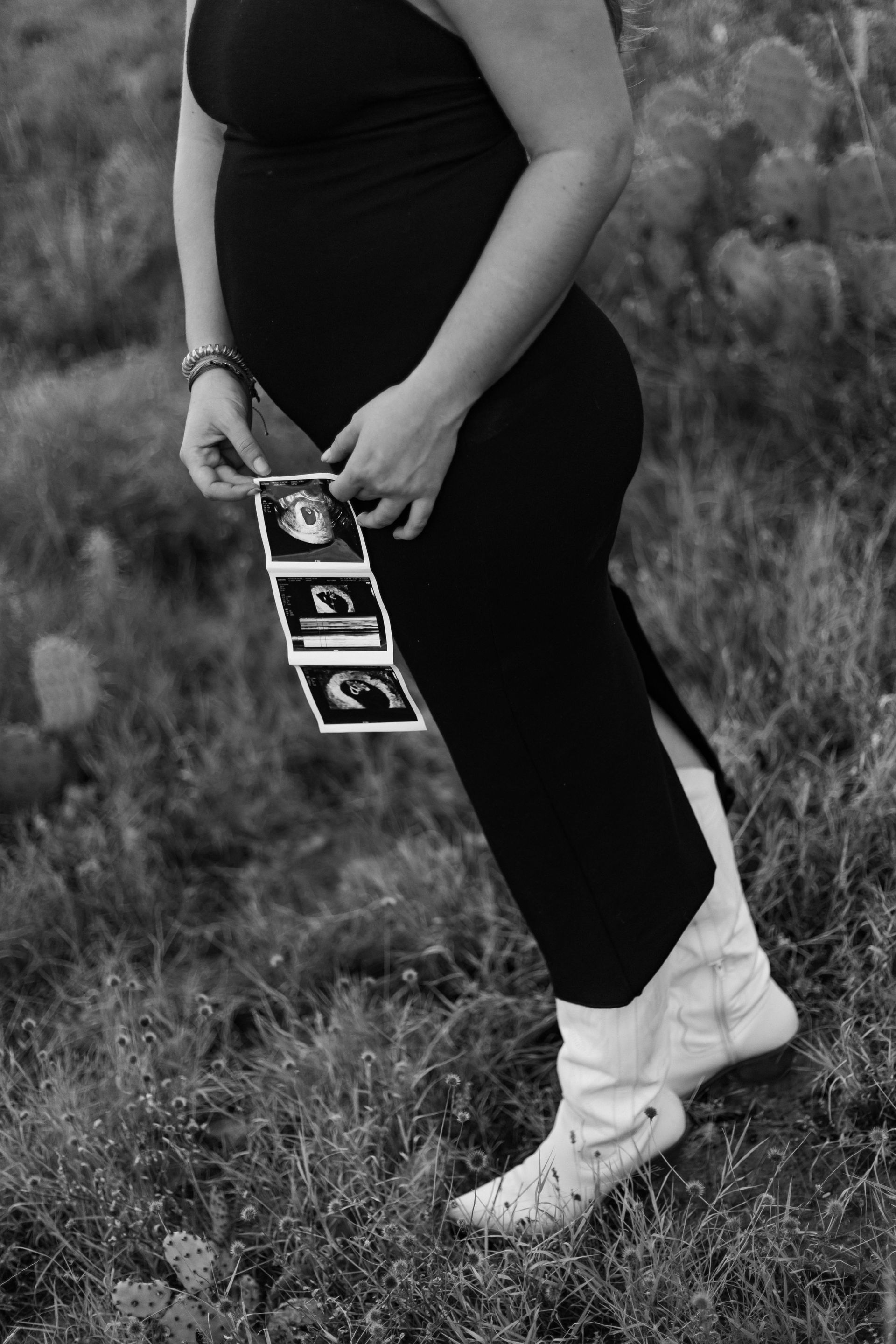 Person standing in a grassy field holding ultrasound photos, wearing a black dress and white boots