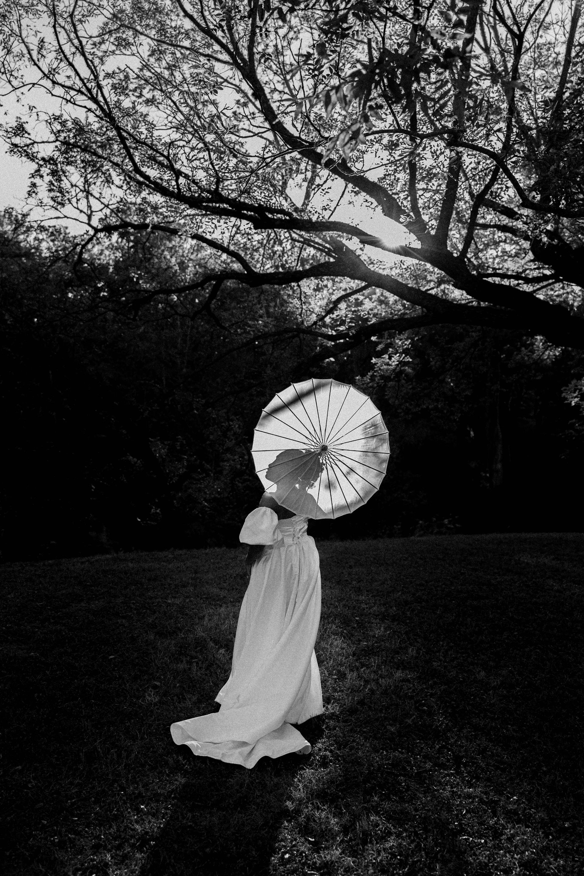 Person in a white dress holding a parasol in a dark wooded field at night.