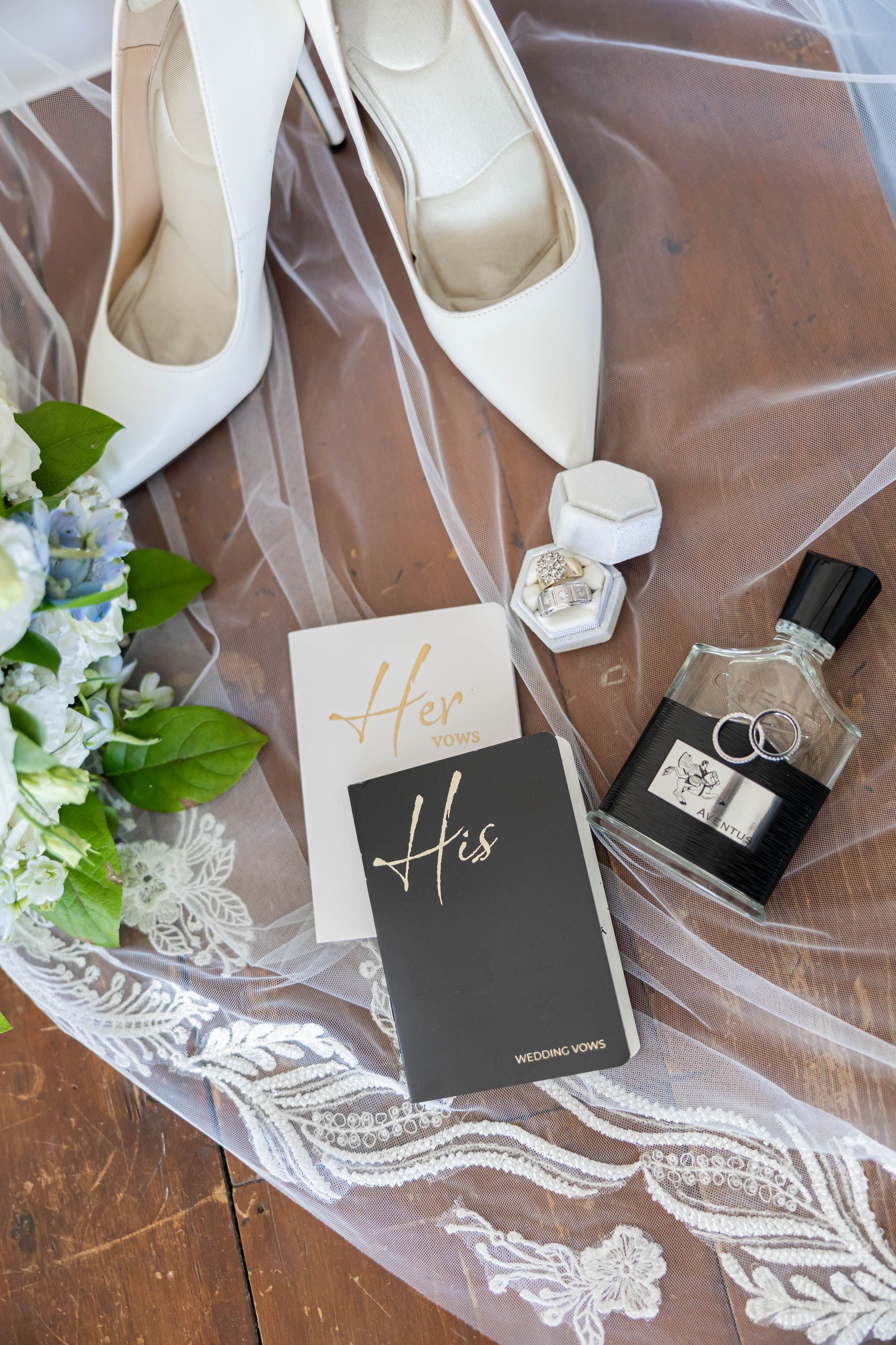 White bridal heels, perfume, rings, and invitation on lace fabric with blue flowers on a wooden floor