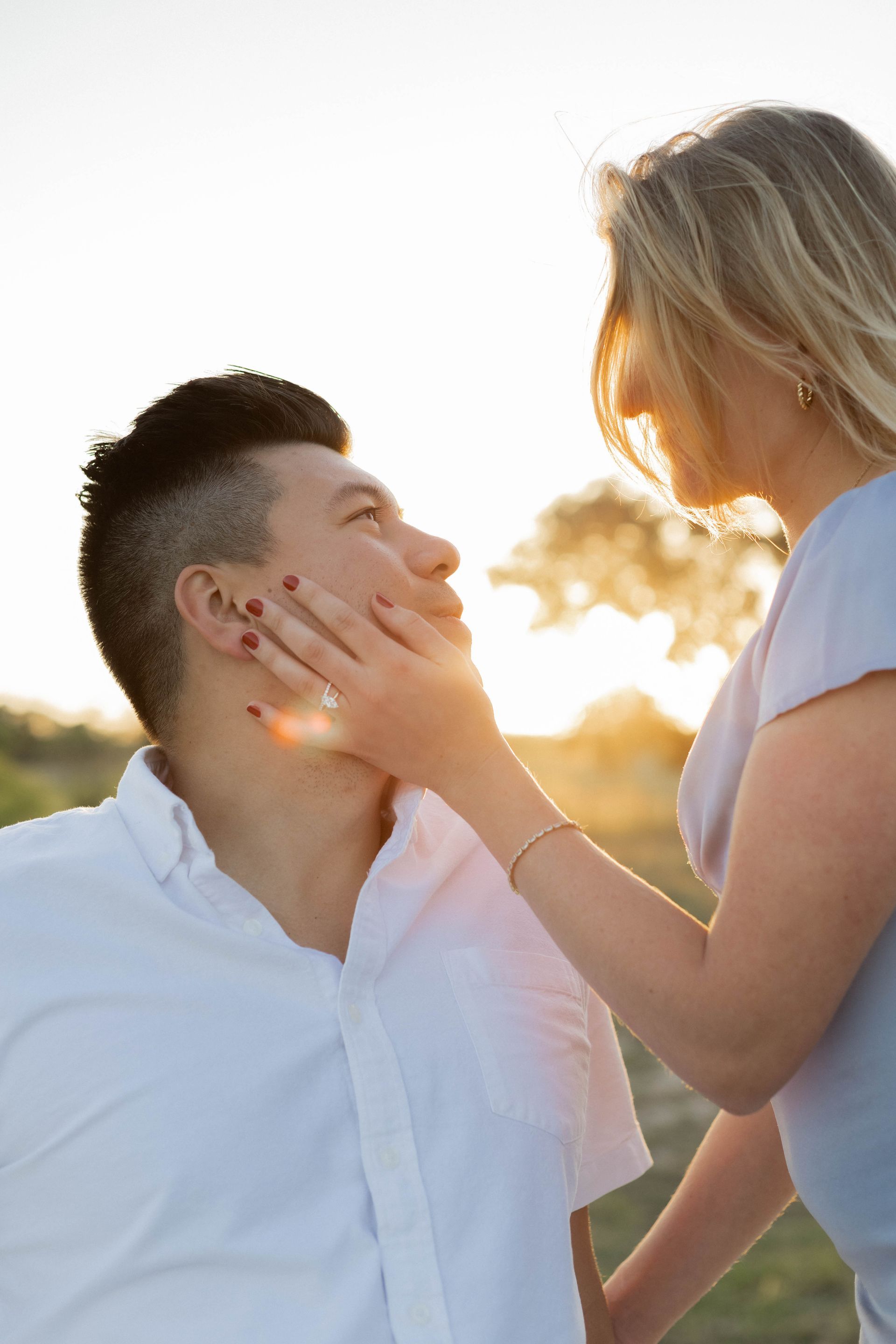 Two people outdoors at sunset, one gently touching the other's face with a warm backlit glow