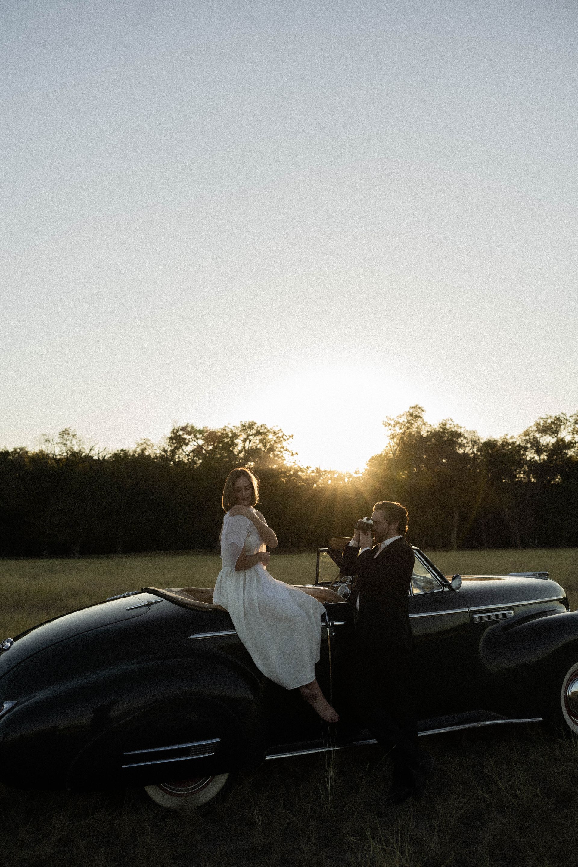 Two people posing on a black vintage car at sunset in a field