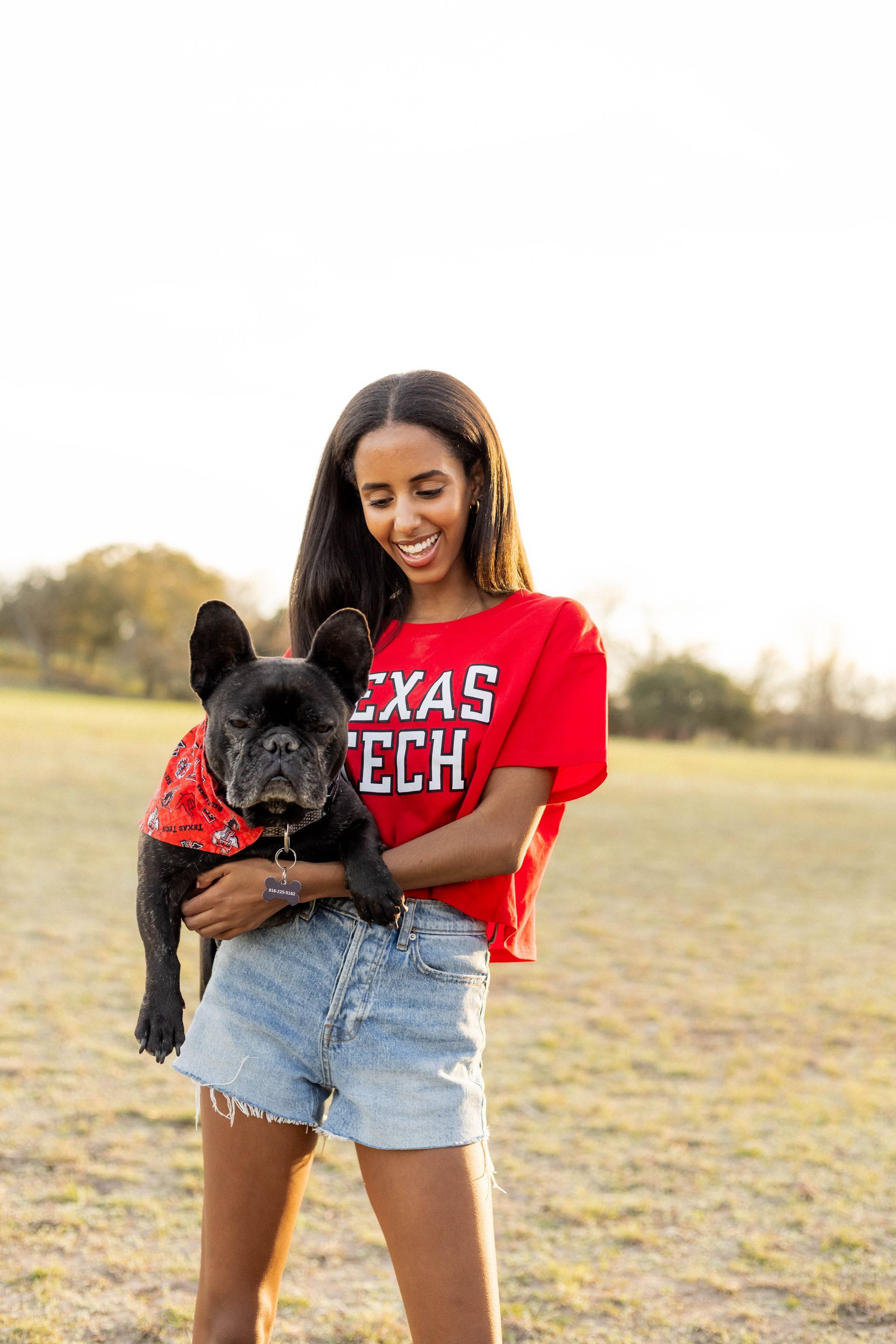 Woman in a red Texas Tech shirt holding a black dog outdoors in a grassy field.