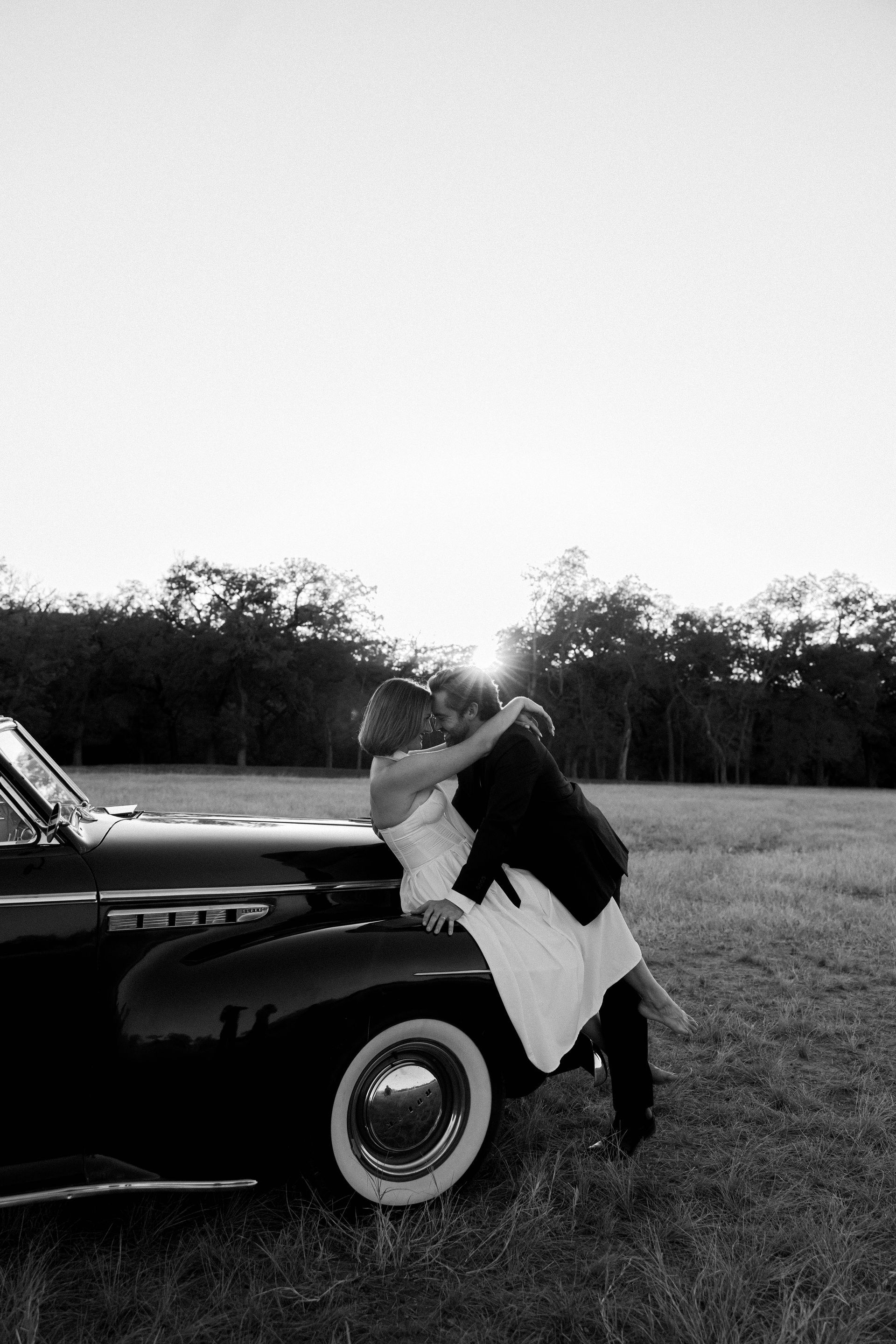 Person leaning on a black vintage car in a grassy field.