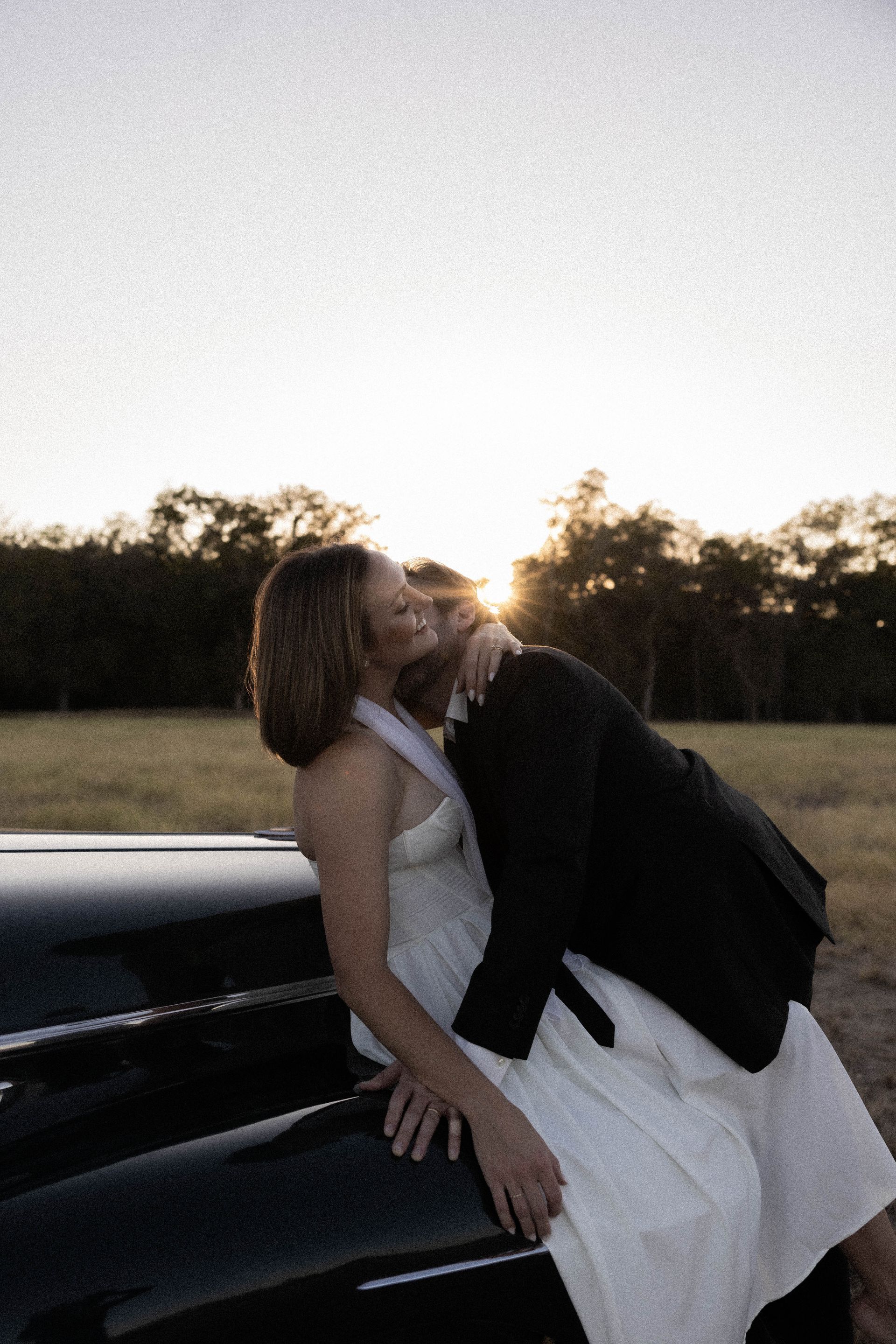Woman in a white dress leaning against a car at sunset in a grassy field