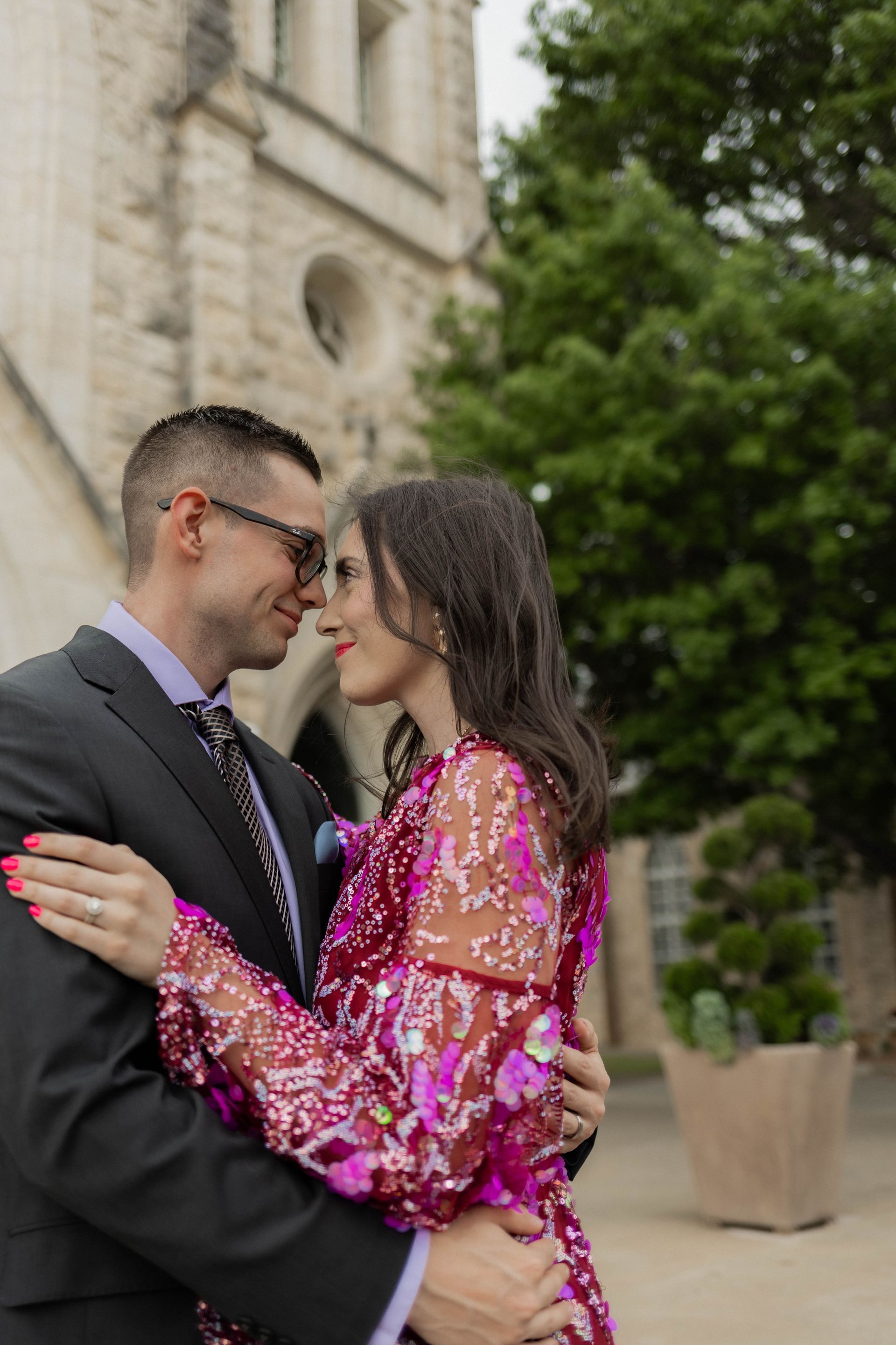 Couple embracing and smiling outside a stone building, the woman in a pink sequined dress