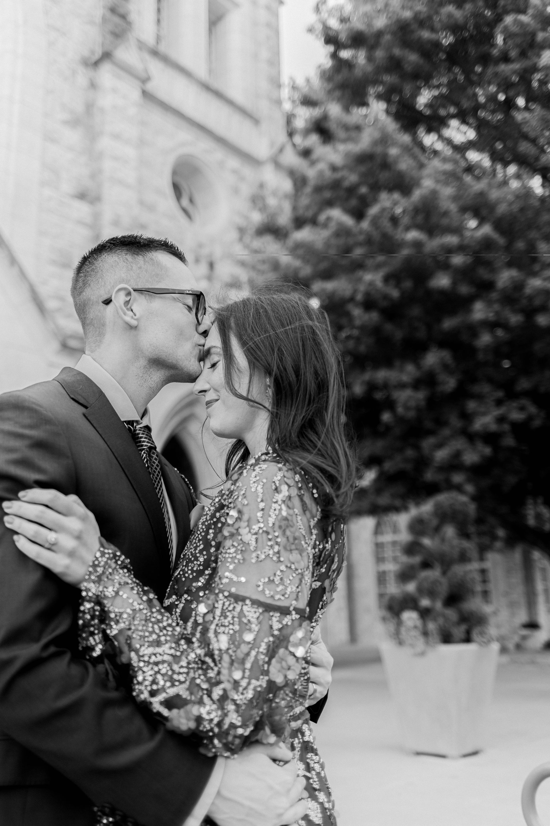Couple embracing and kissing outdoors near a building, with trees in the background.
