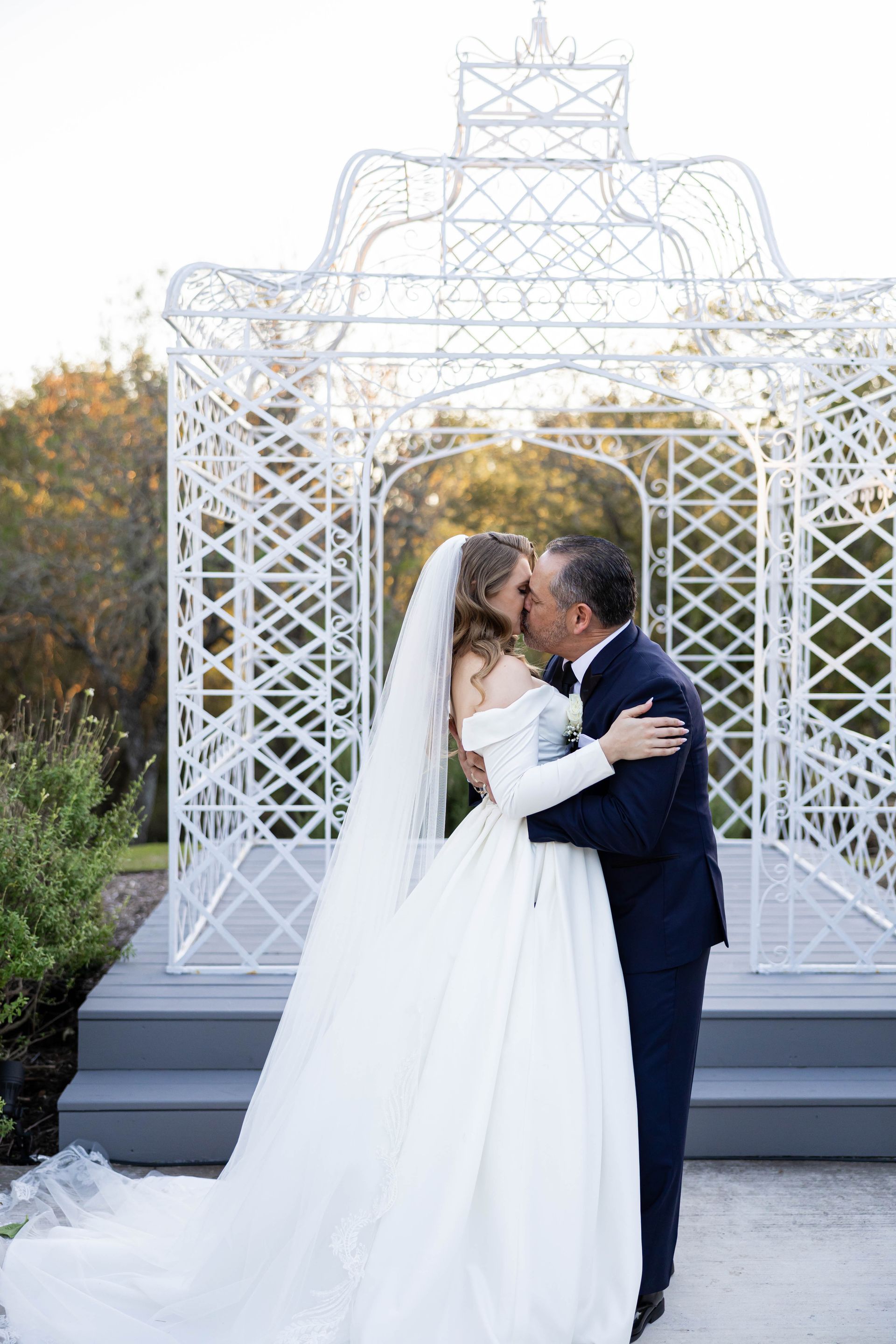 Bride and groom kissing beneath a white decorative arch outdoors