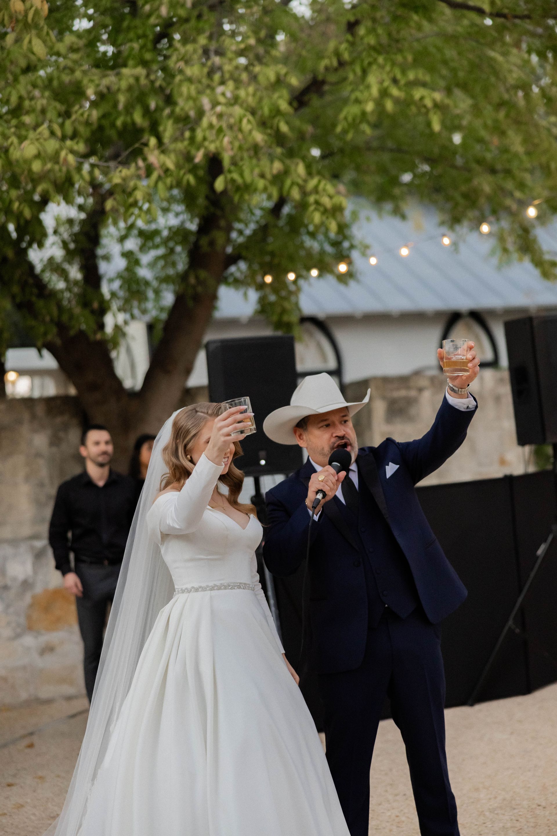 Wedding couple in white gown and dark suit, raising drinks outdoors under a tree at dusk