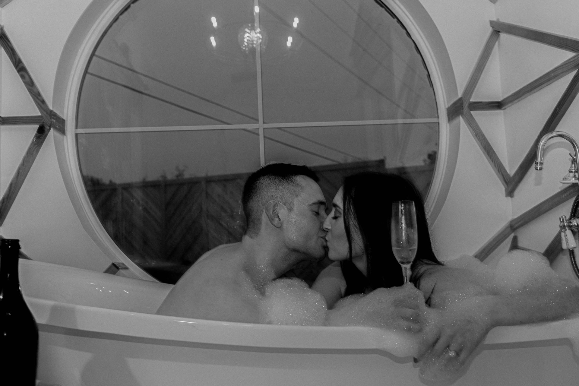 Couple kissing in a bathtub by a round window, with a champagne flute nearby.