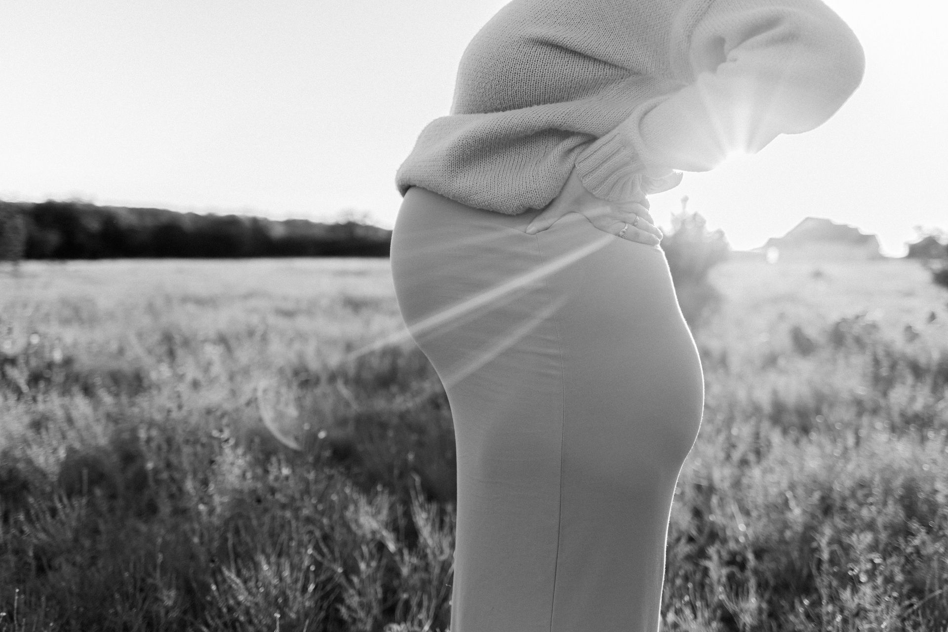 Pregnant person in a field, backlit by the sun, holding their belly in black and white.