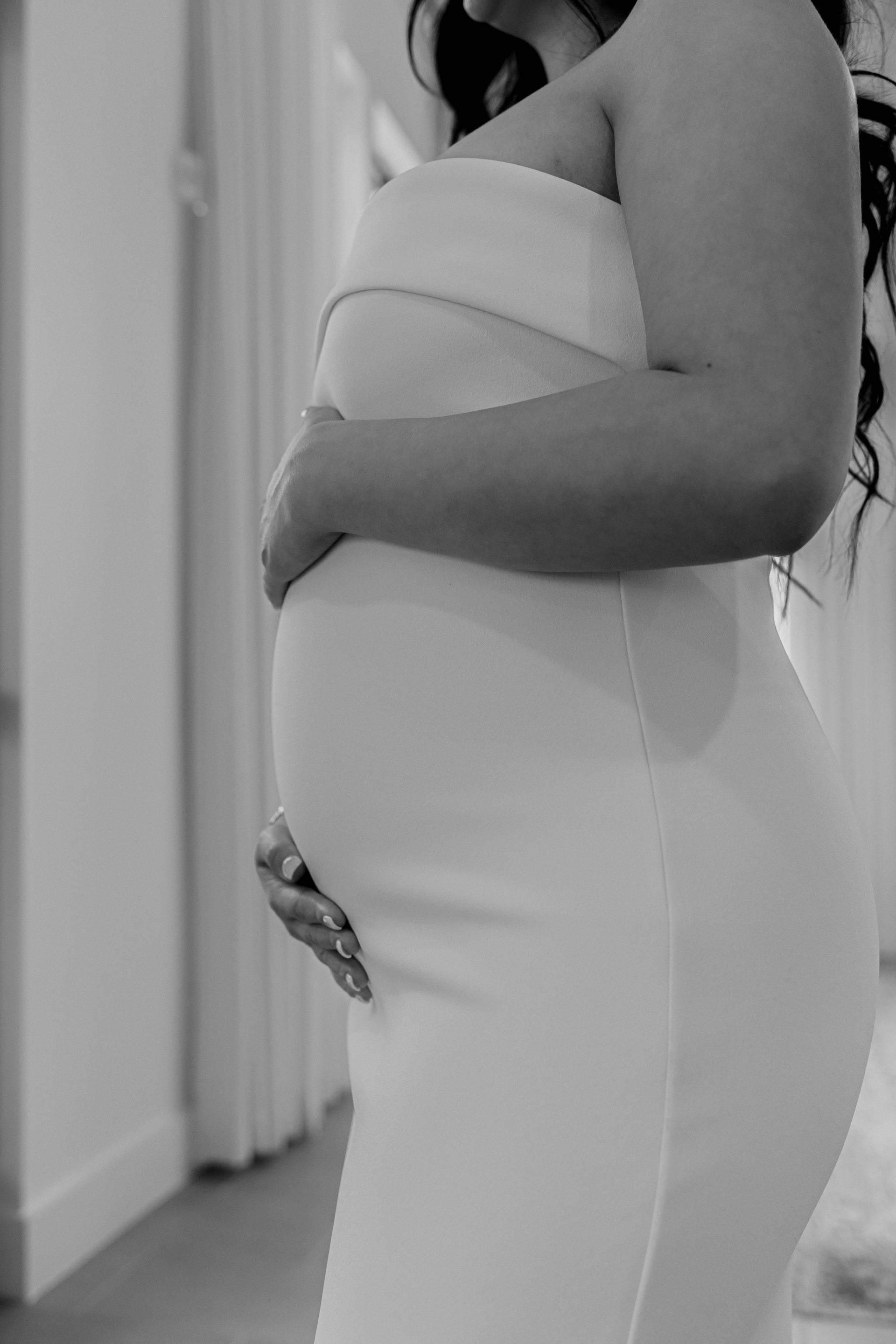 Side view of a person in a white dress, holding their pregnant belly indoors.