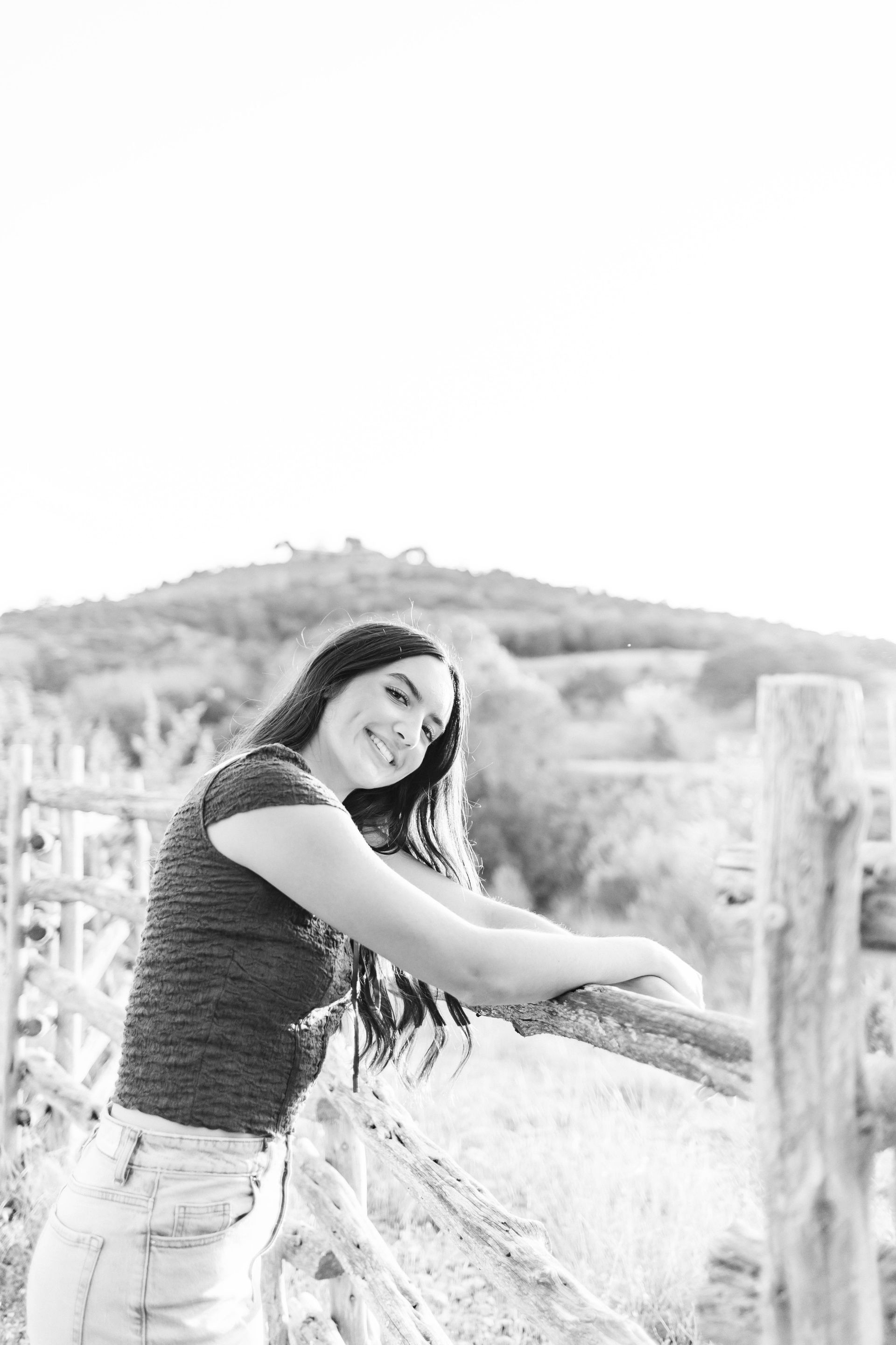Woman smiling and leaning on a fence in a bright outdoor field with hills in the background