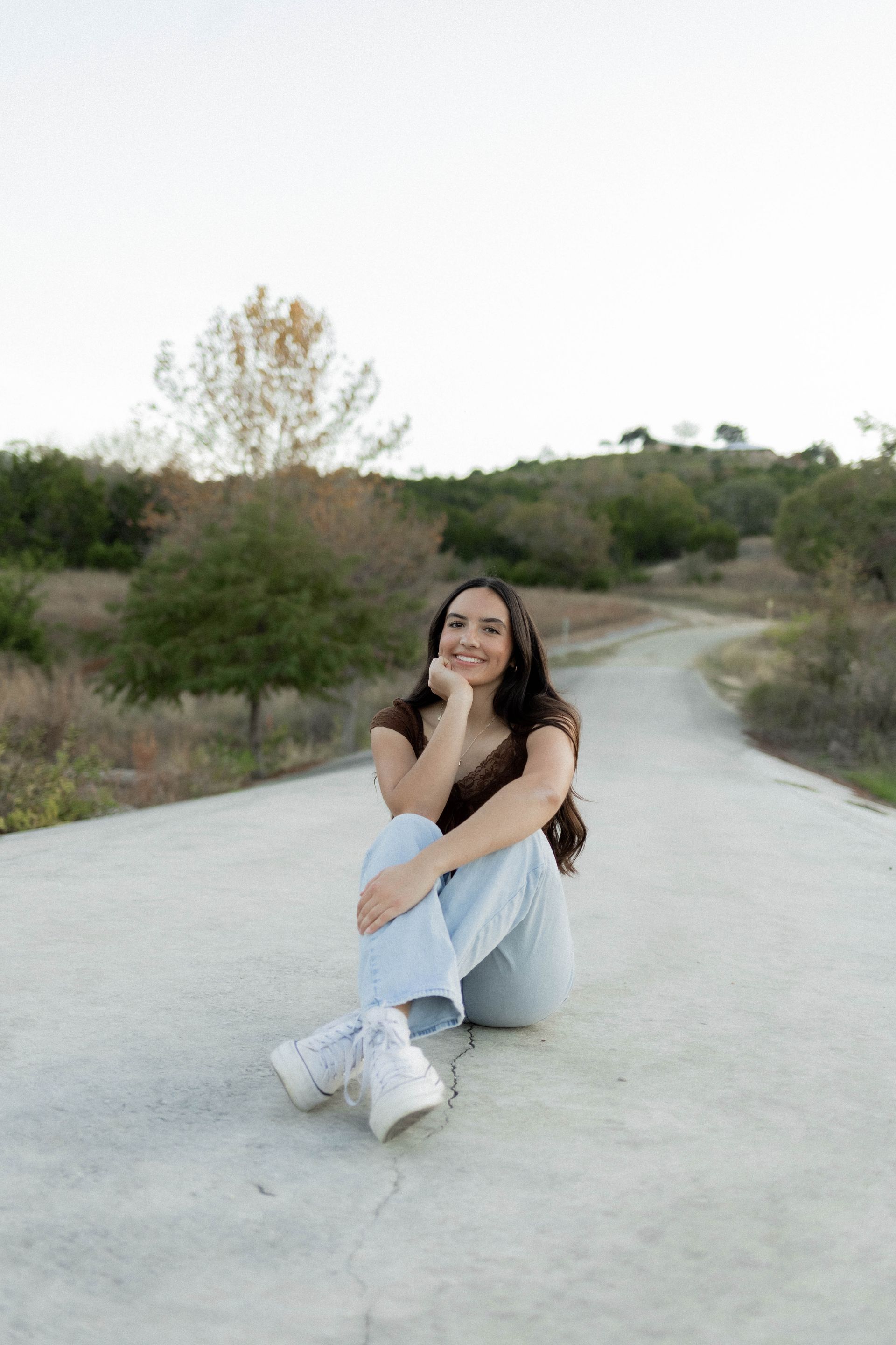 Person sitting on a winding road in a grassy landscape, smiling with a hand on their face.