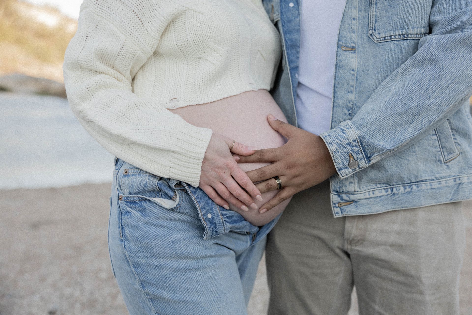 Two people standing close together, holding hands over a pregnant belly outdoors.