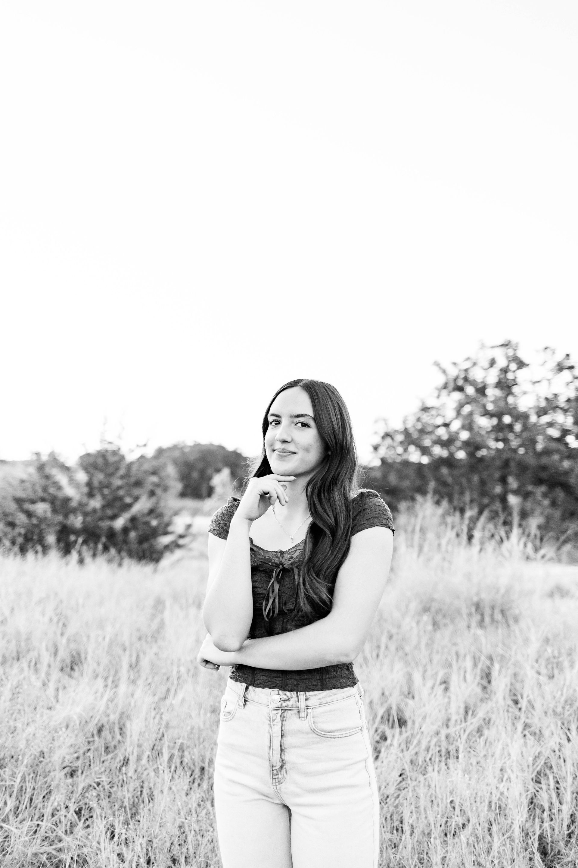 Smiling woman standing in tall grass with one hand on her chin, outdoors on a cloudy day