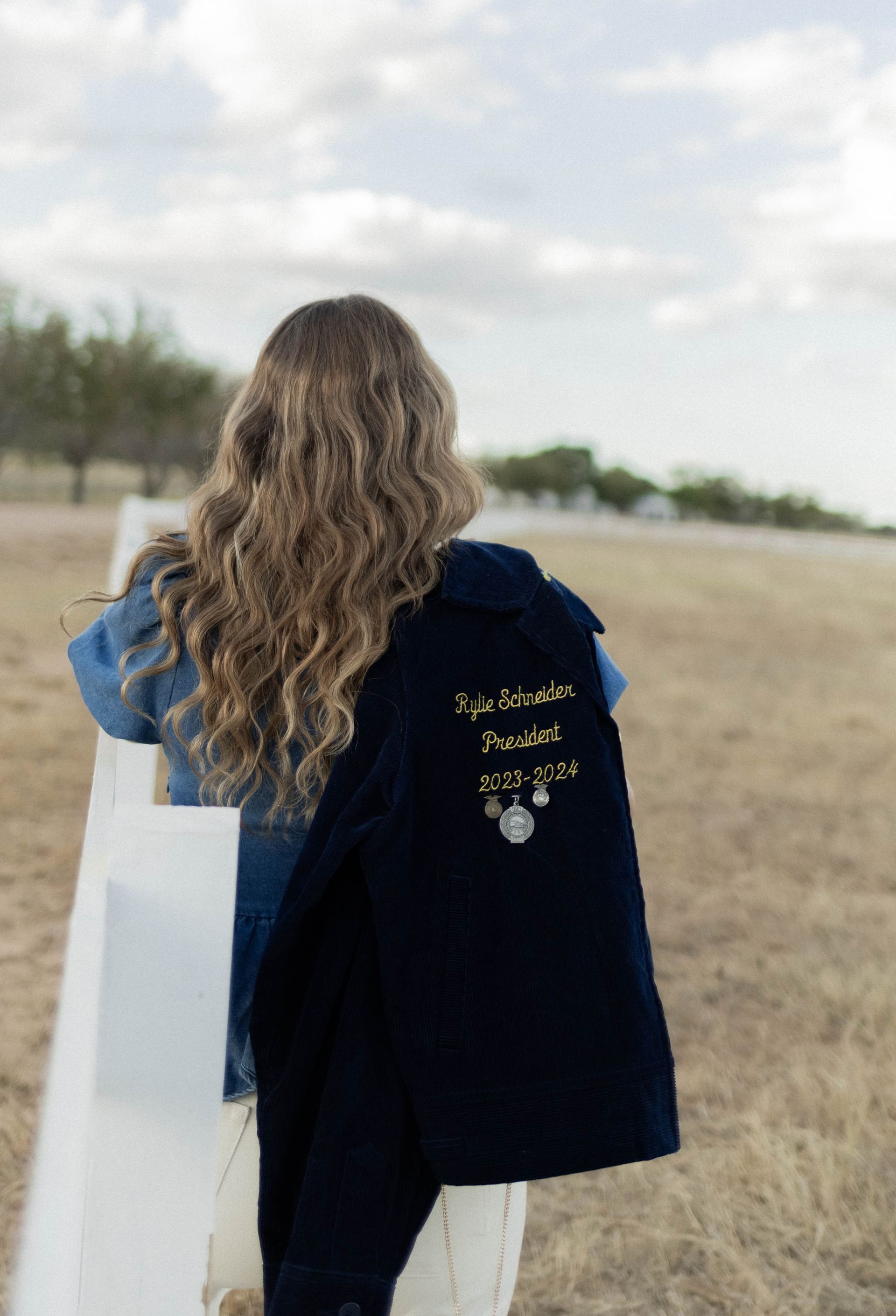 Person walking in a grassy field with a blue tote bag and dark jacket over one shoulder.
