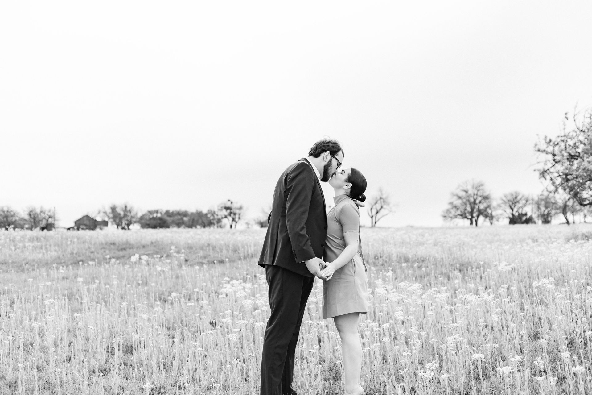 Couple kissing in a field, holding hands amid tall grass under a bright sky