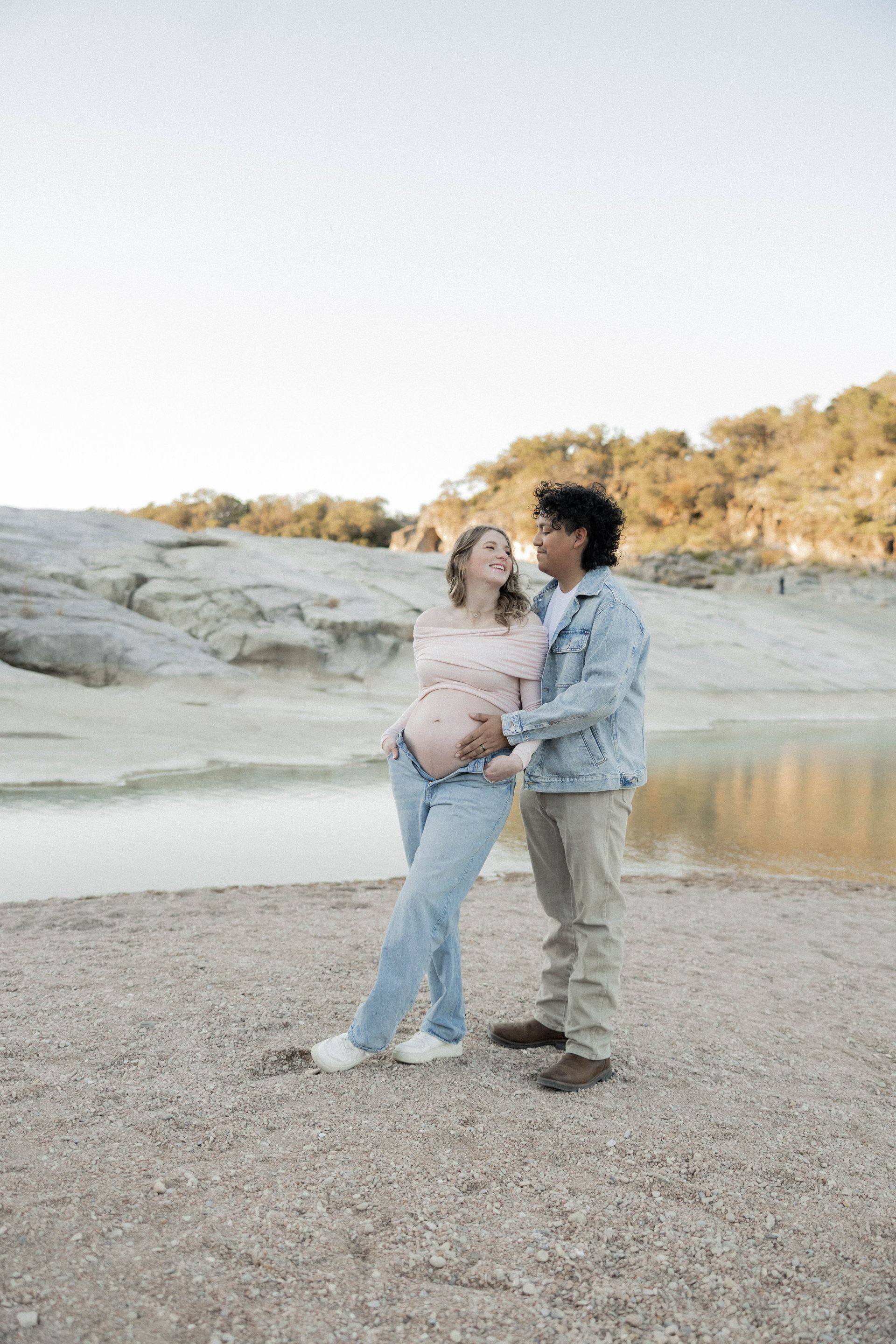 Couple standing close together on a rocky beach at sunset, with calm water and cliffs in the background