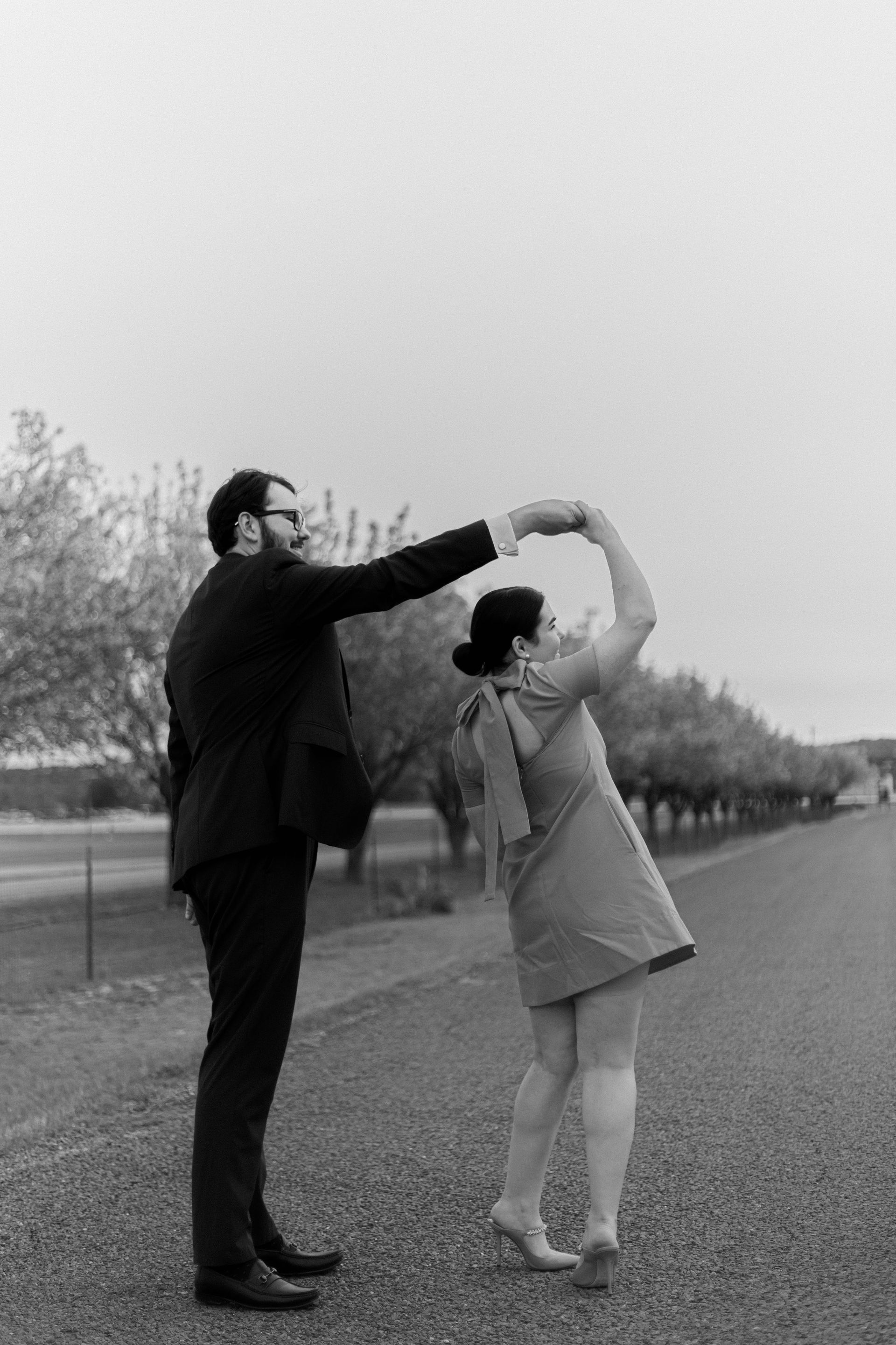 Two people outdoors on a road, one leading the other in a dance pose with arms raised.