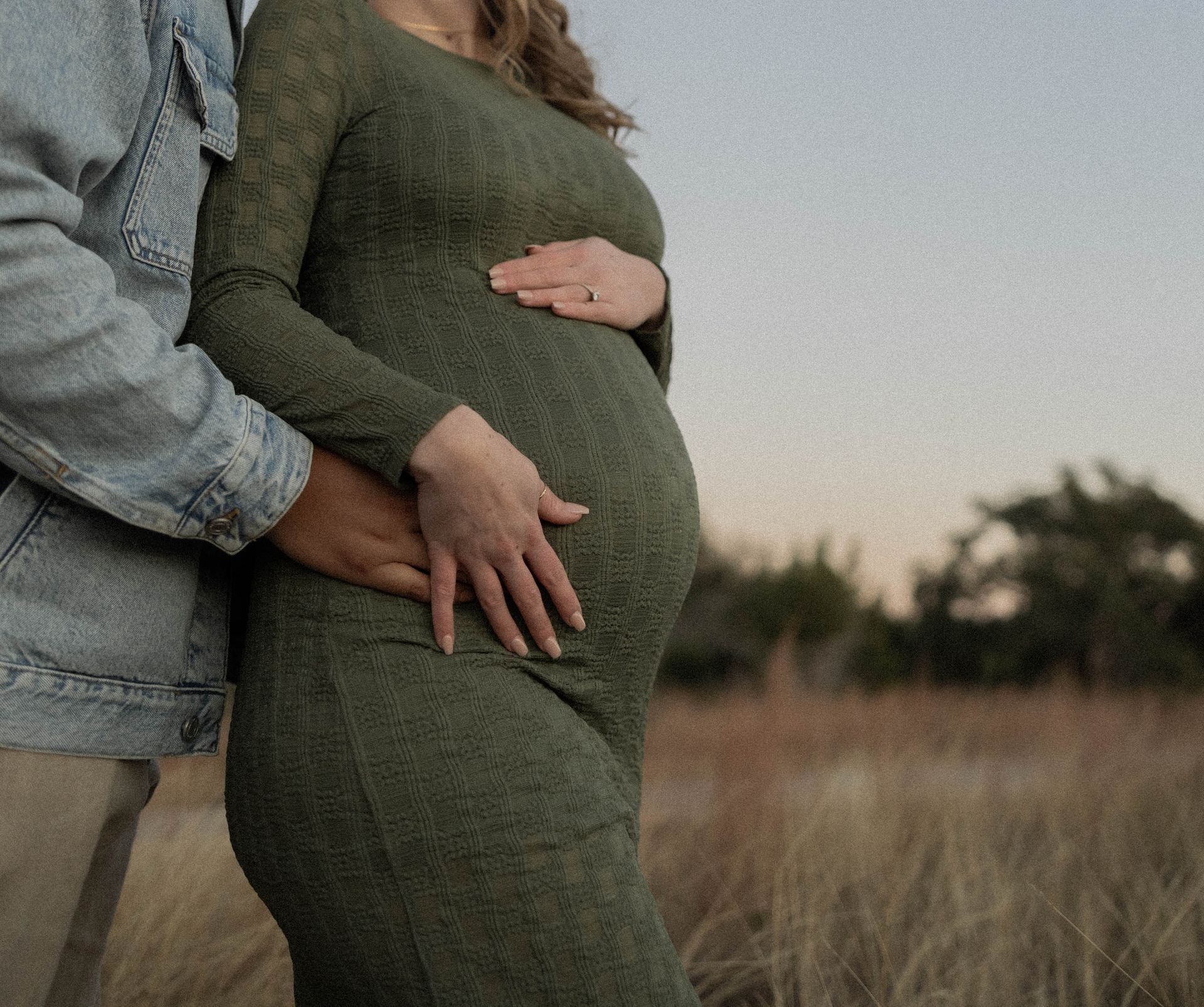 Couple embracing a pregnant belly in a grassy field at dusk