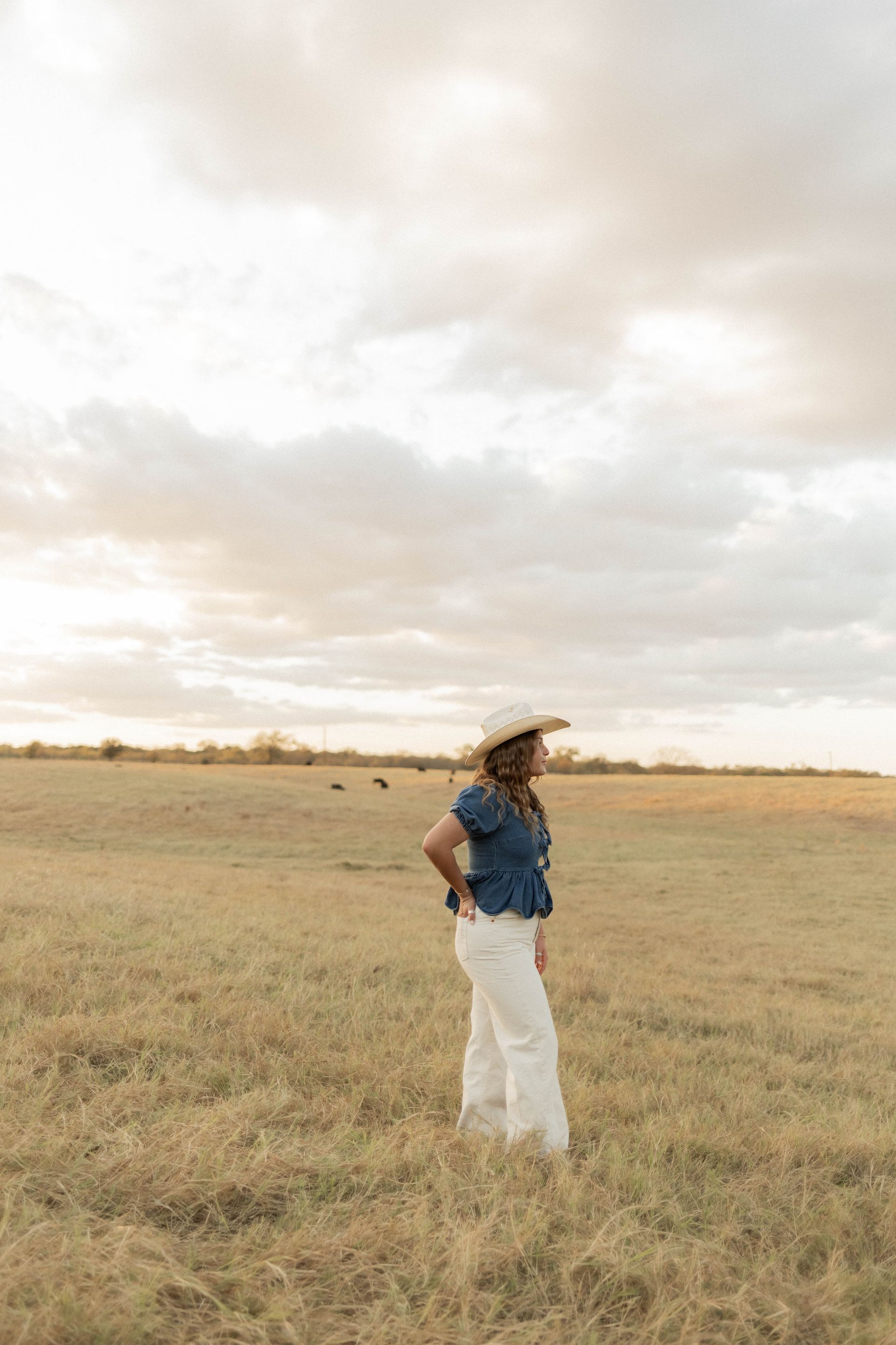 Person in a blue shirt and white pants standing in a grassy field under a cloudy sky.
