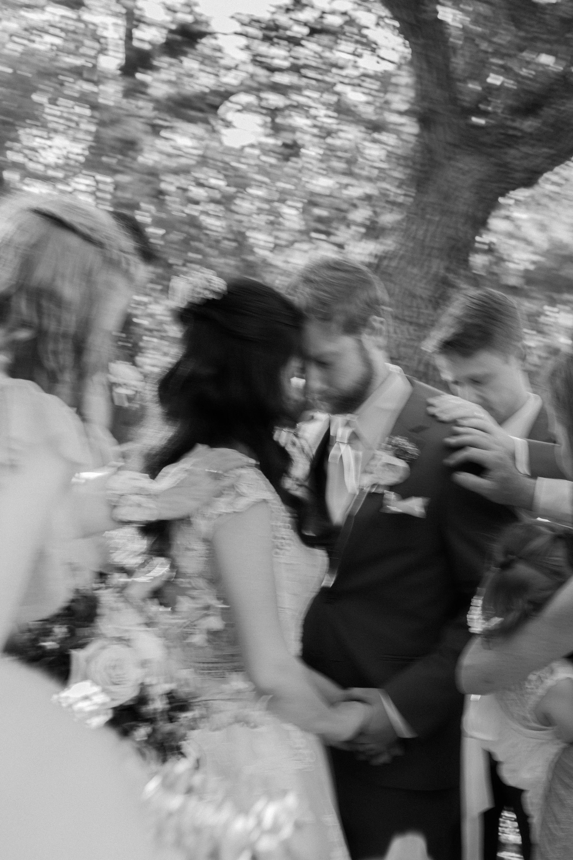 Bride and groom embracing outdoors at a wedding ceremony, with guests nearby and trees in the background