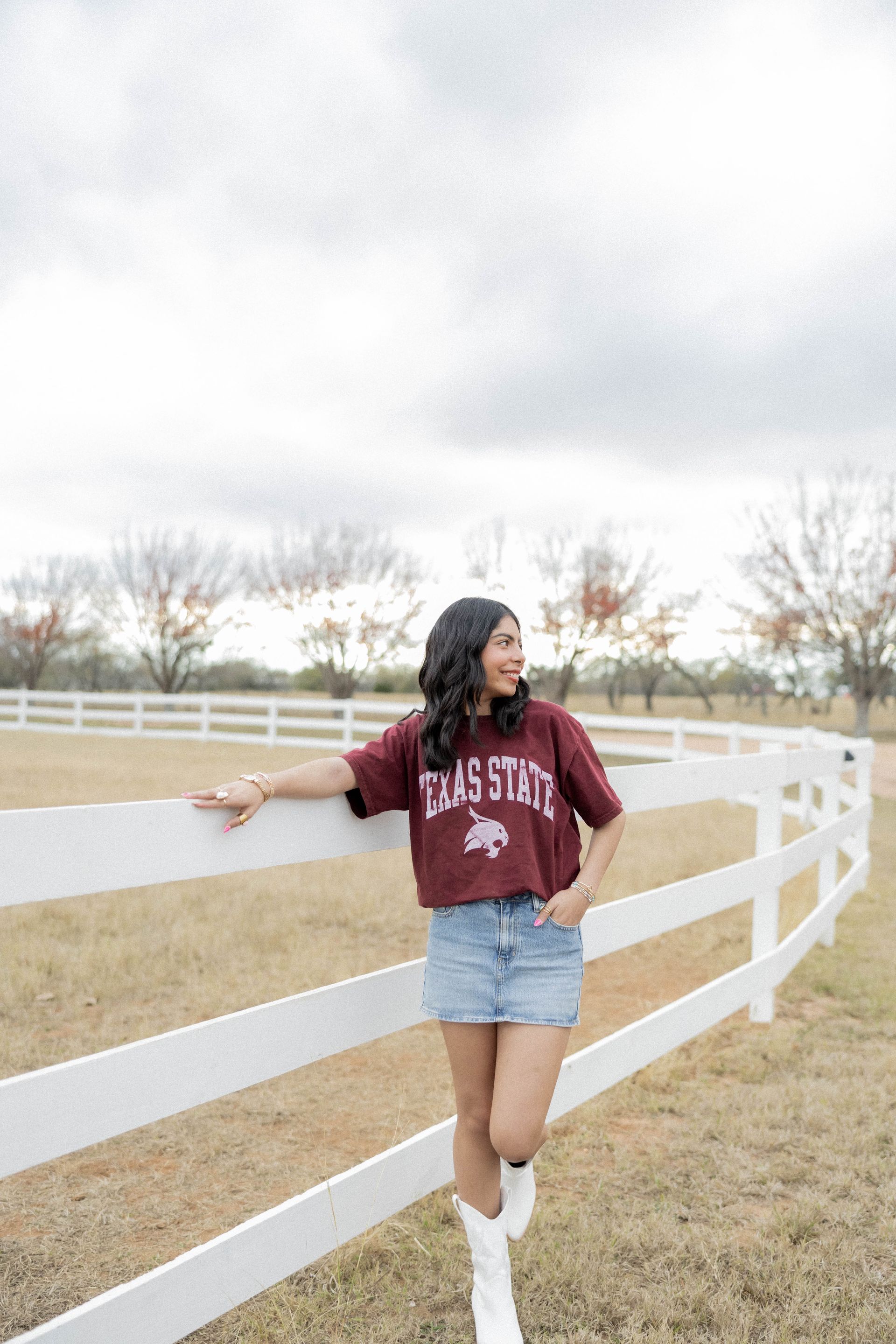 Person leaning on a white fence in a grassy field, wearing a maroon sweatshirt, denim shorts, and white boots.