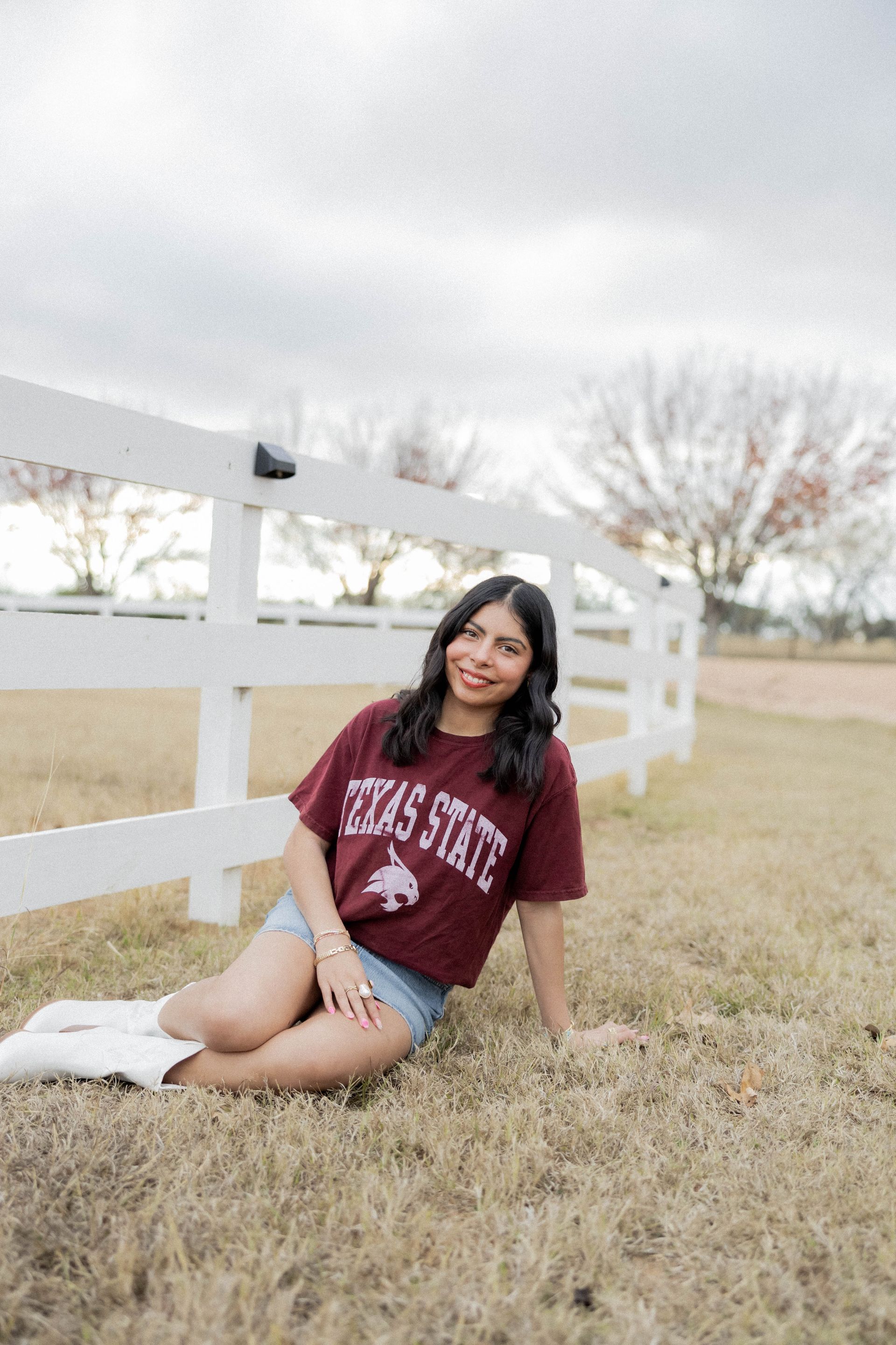 Woman sitting in grass beside a white fence, wearing a maroon sweatshirt and smiling at the camera.