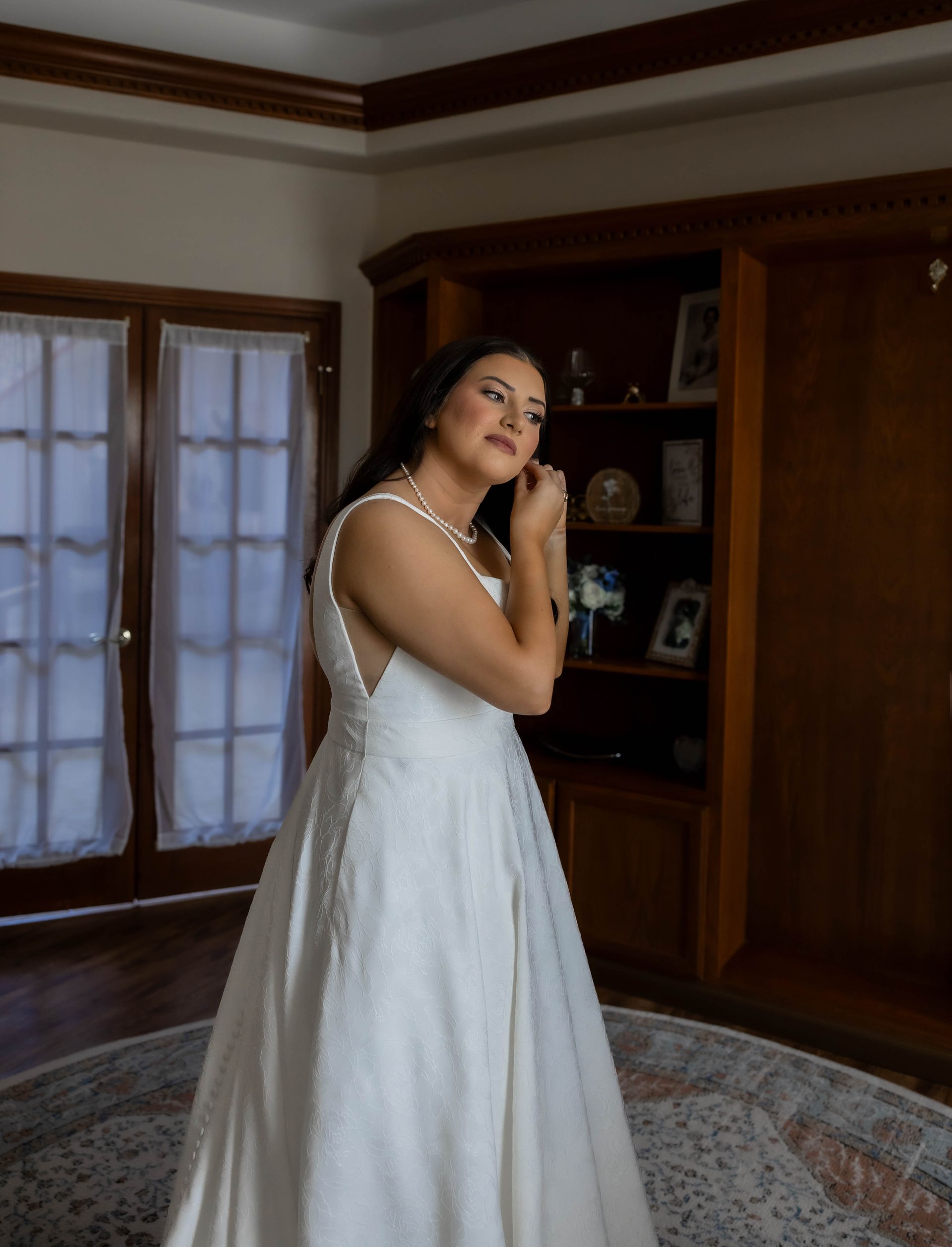 Woman in a white gown standing indoors beside a wooden bookcase and windows, adjusting her hair.