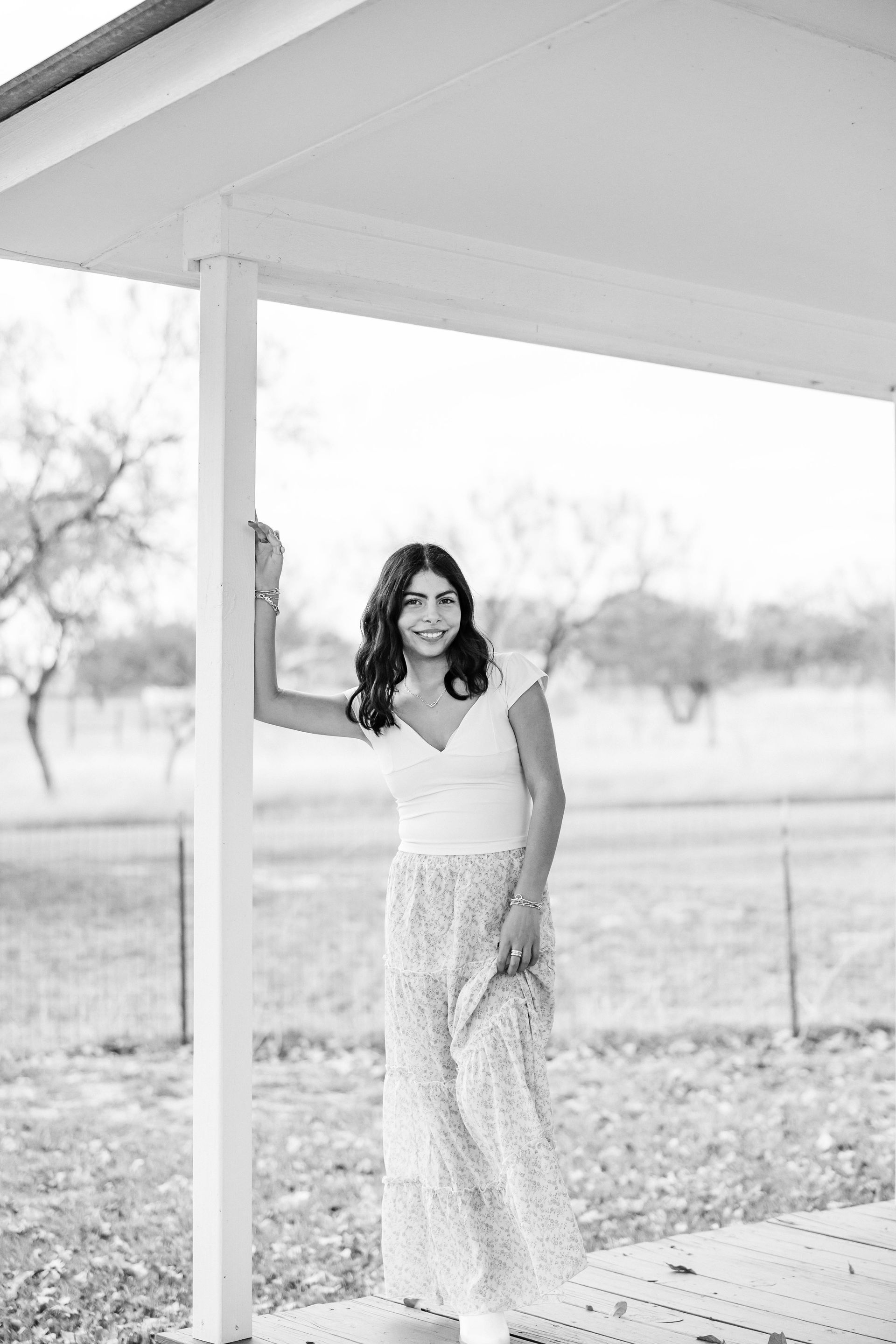 Woman in a white dress standing by a porch post in a field, smiling at the camera.