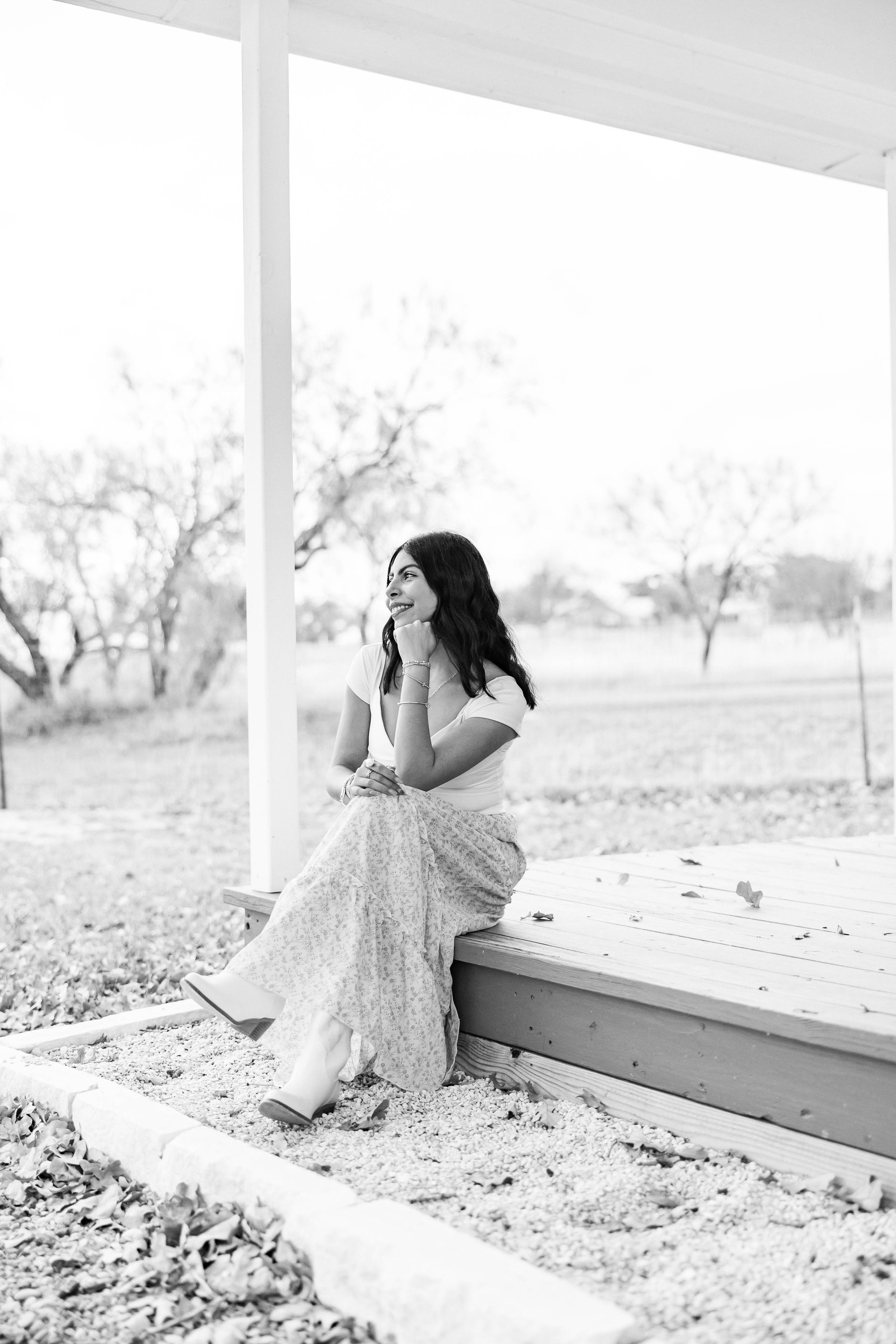 Woman sitting on a porch in a white dress, looking thoughtful in a bright outdoor garden setting