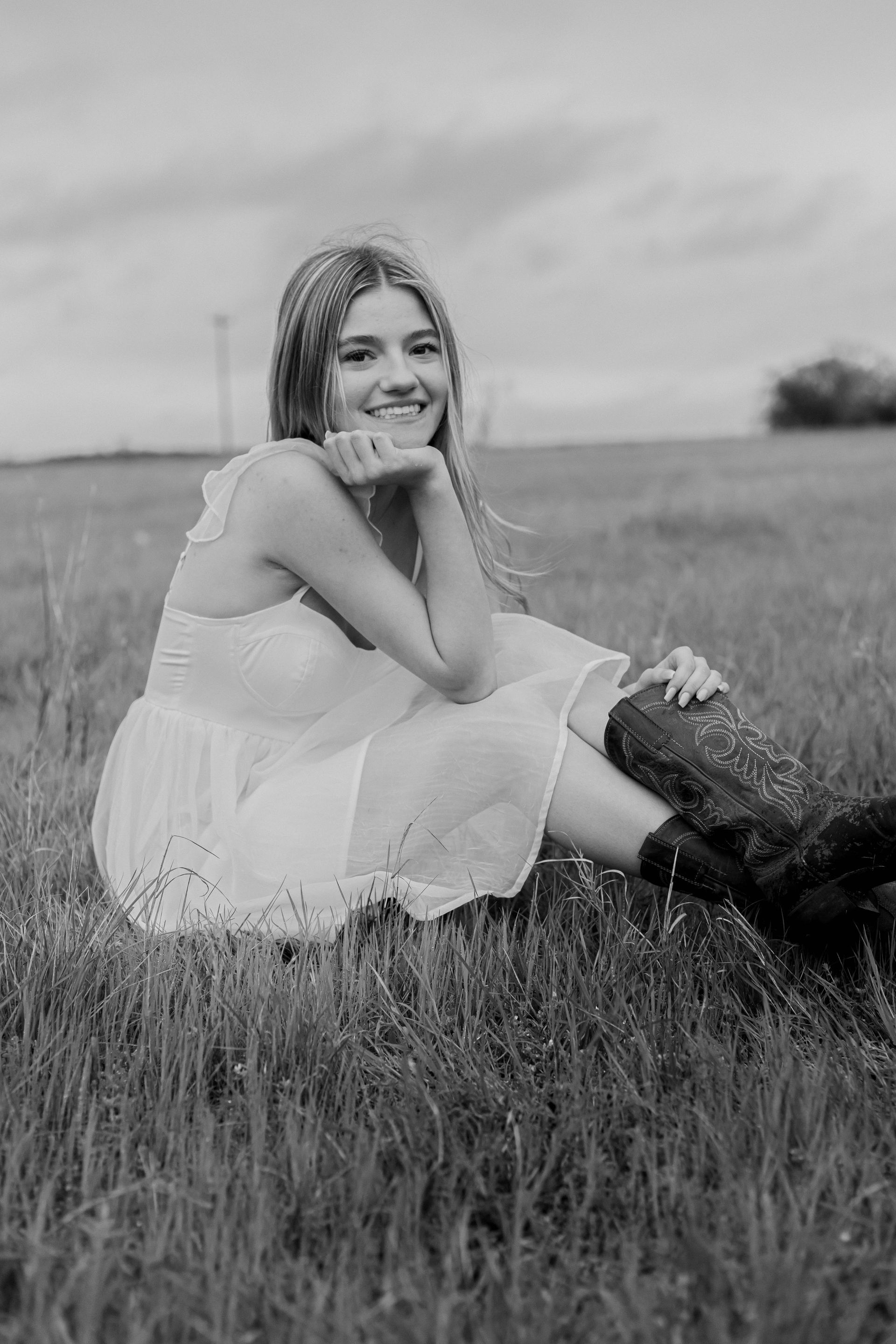 Young woman in a white dress sitting in a grassy field, smiling at the camera