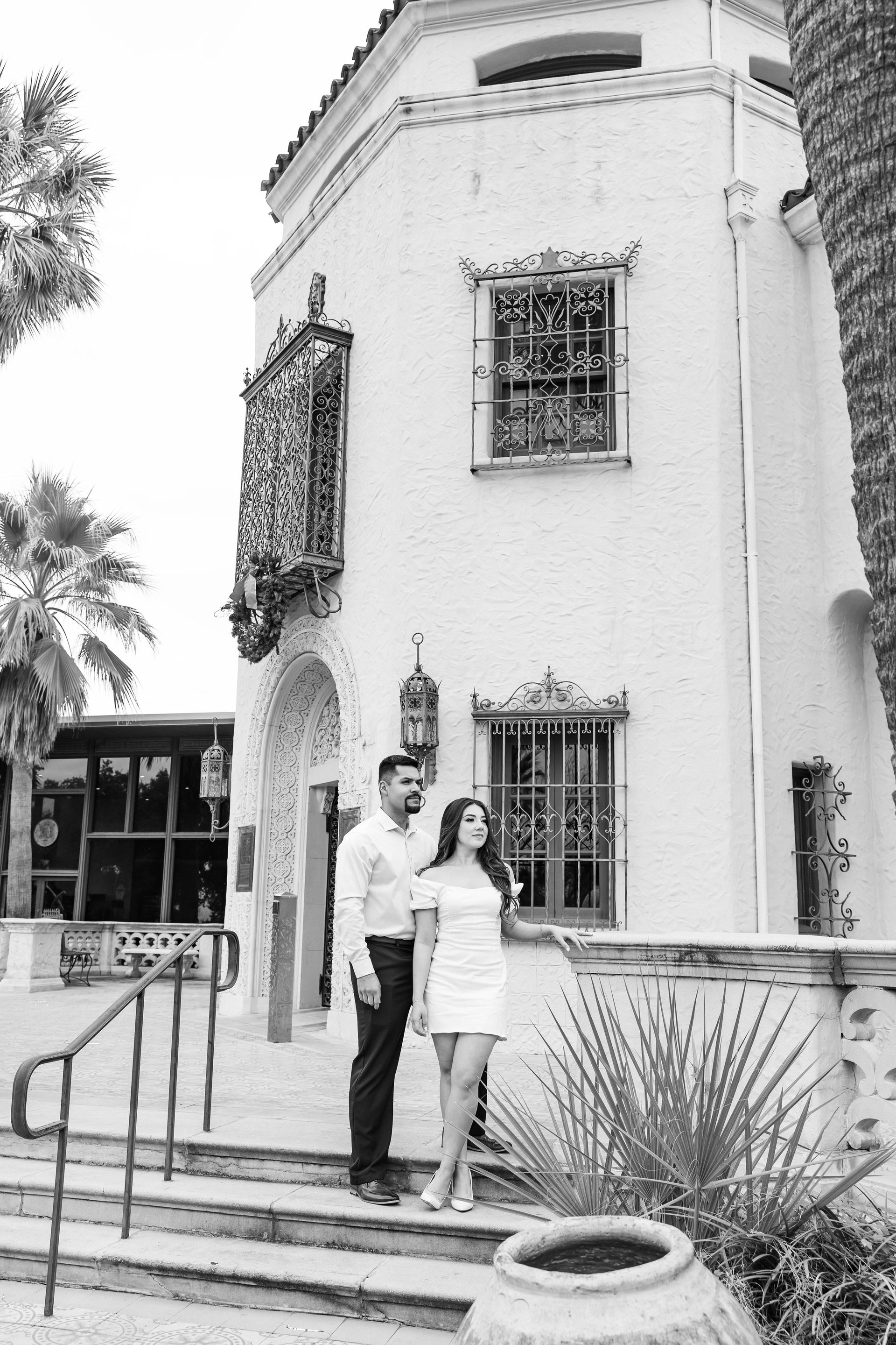 Couple standing on steps outside a white stucco building with arched windows and potted plants